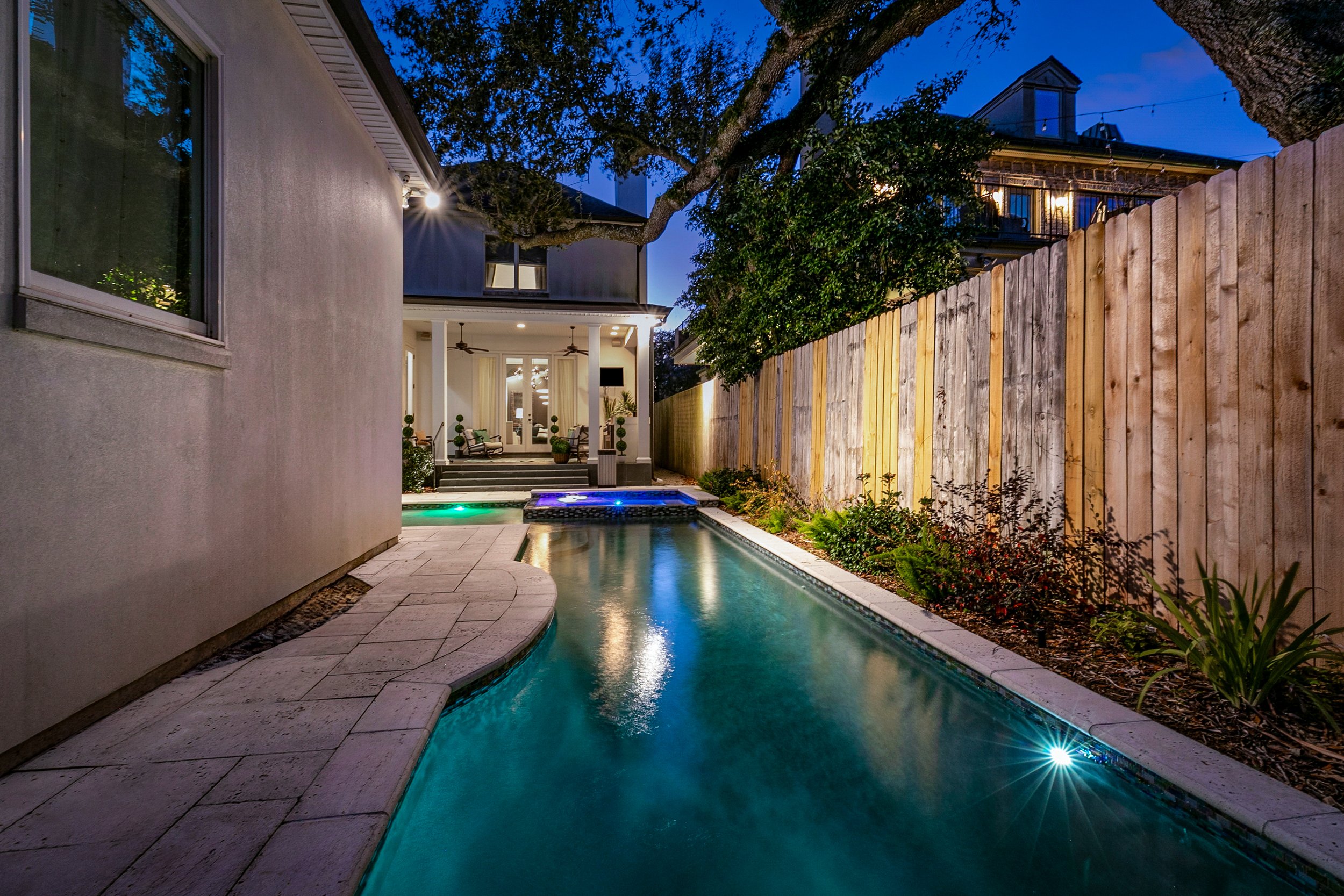 Luxury Listing pictures with Aimee (Amay) Curole, Top Realtor in Metairie, 23 Realty. Backyard with a swimming pool, fenced with a wooden fence, adjacent to a house with outdoor seating, trees, and evening lighting.