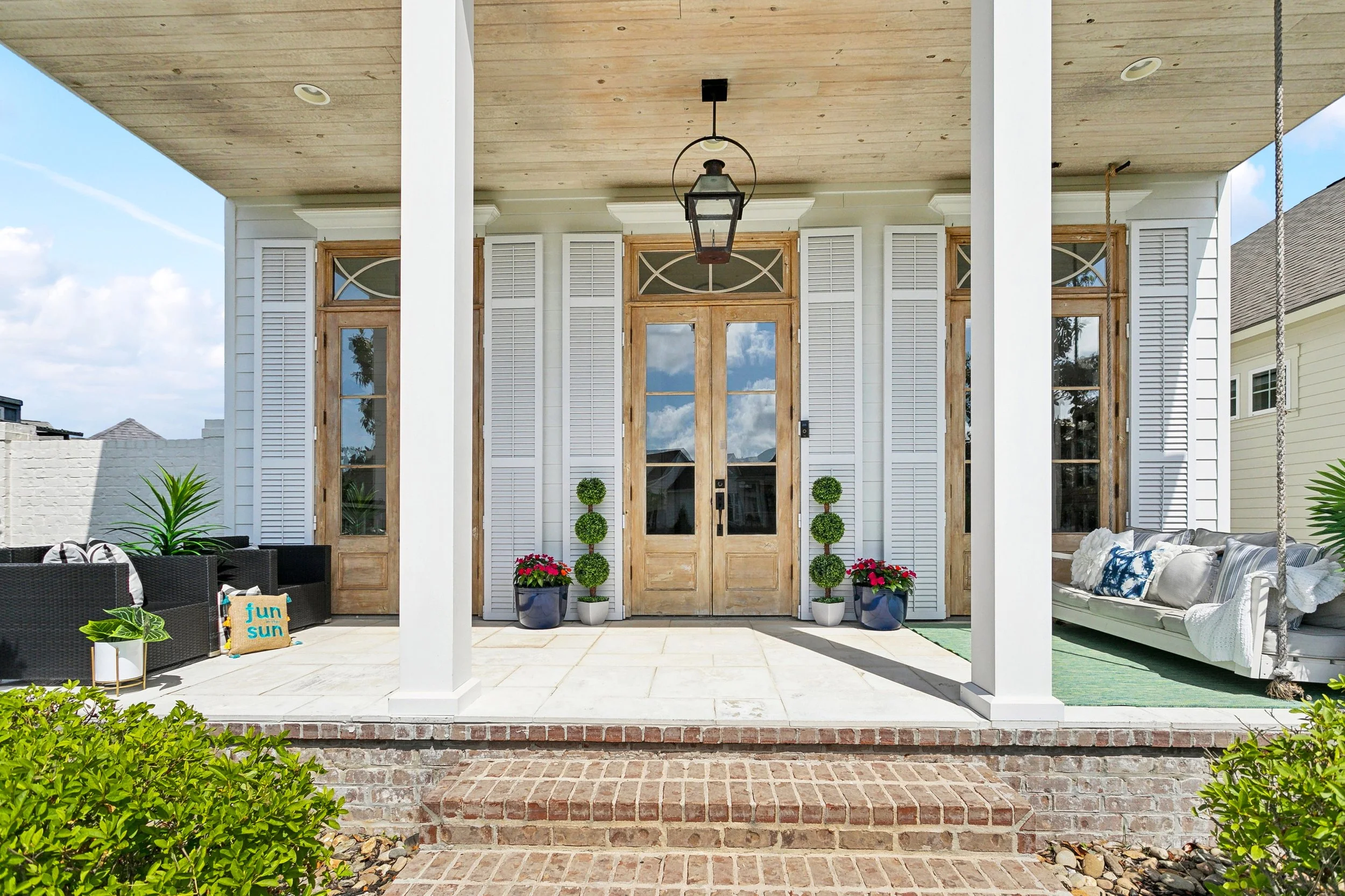 Front porch of a house with wooden double doors, potted topiary trees, and decorative shutters. Outdoor seating with pillows and plants, a hanging lantern, brick stairs, and a small garden area.