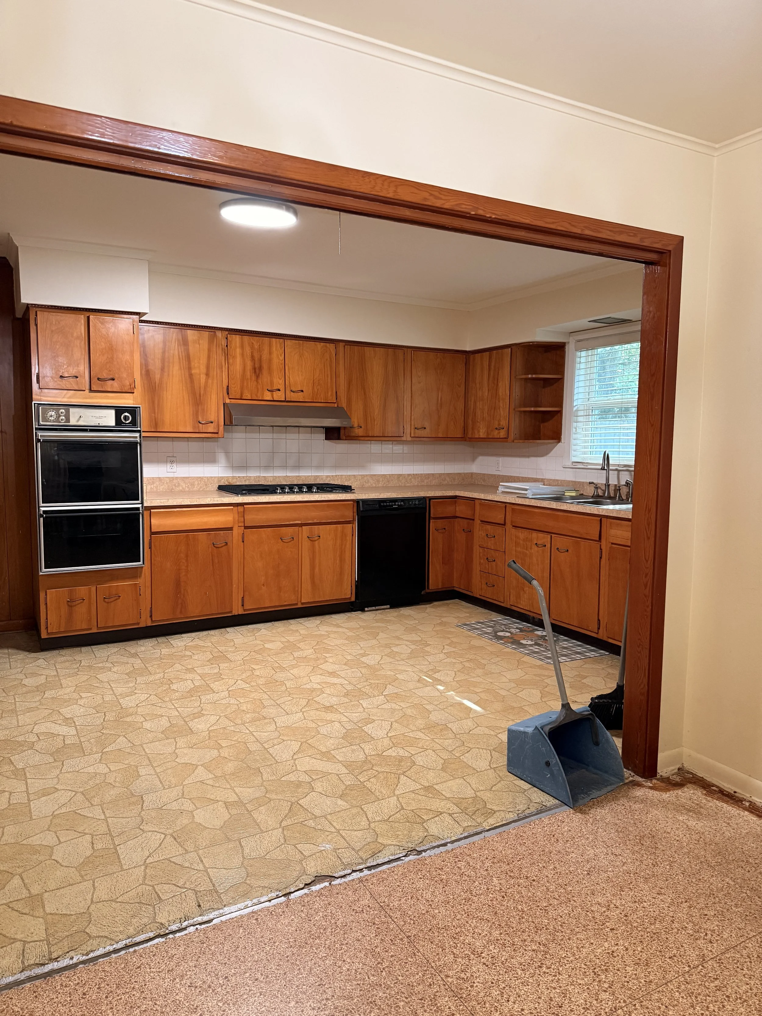 View through a doorway into a kitchen with wooden cabinets, a window with blinds, and a countertop with a sink. A dustpan and broom are placed on the floor near the doorway, which is currently under renovation with a section of the lower floor removed.
