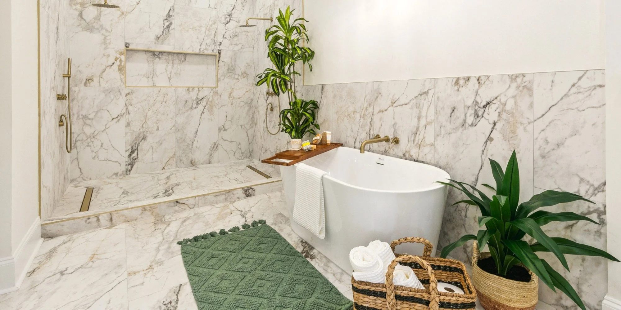 Modern bathroom with a marble shower area, a freestanding bathtub, and green plants. The shower has gold fixtures and a built-in shelf. A wooden tray holds toiletries on the bathtub. A green bath mat and a woven basket with rolled white towels are on the marble floor.