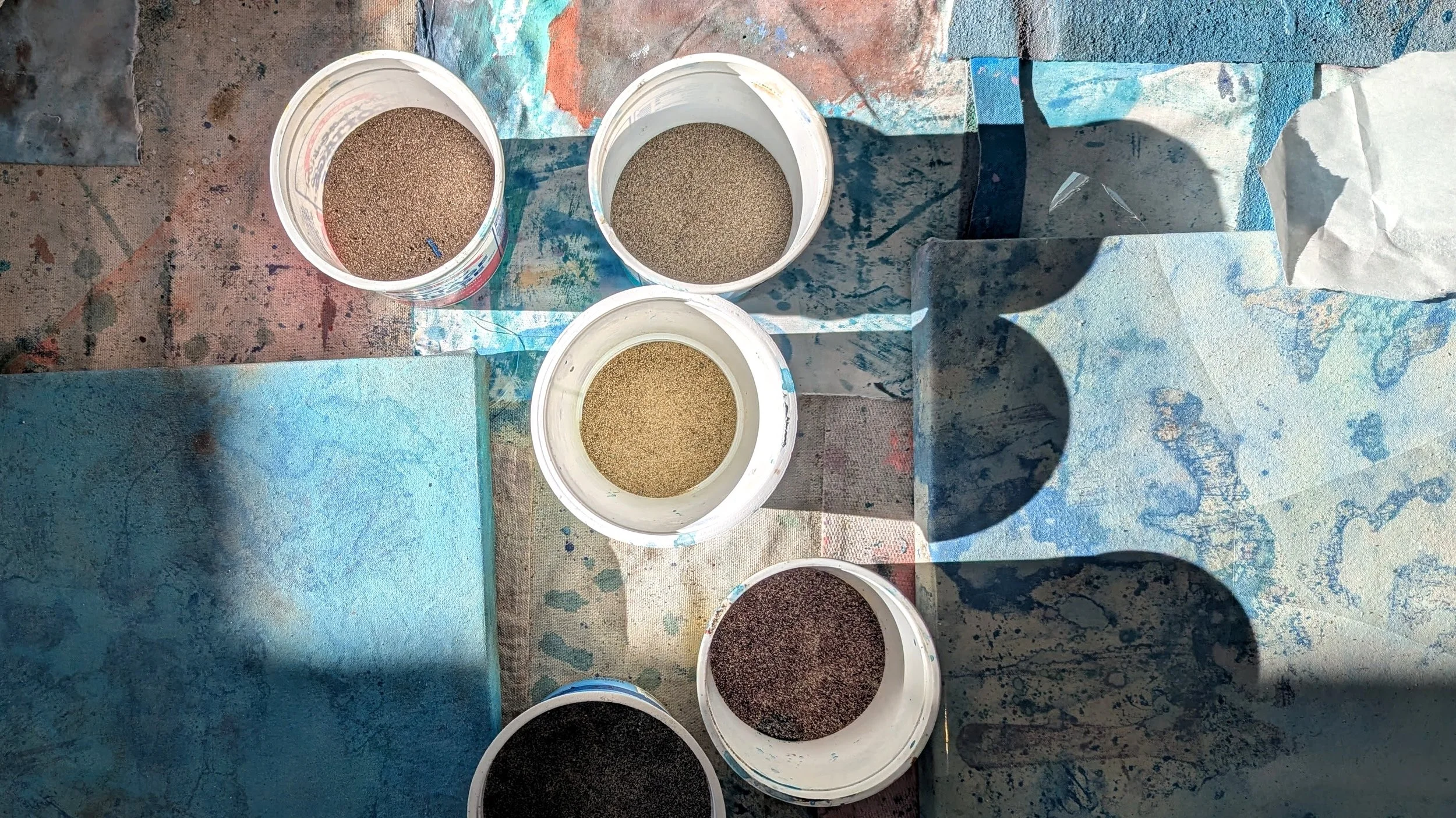 A top-down view of five open containers filled with different colored sand on a table covered with paint splatters, with some blue-painted canvases nearby.