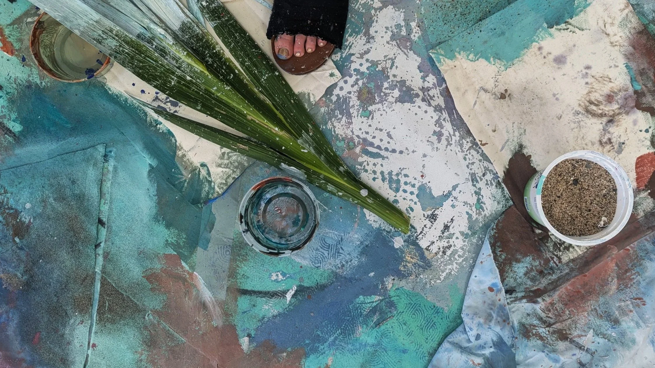 An artist's workspace with abstract painted fabric or paper, a large green leaf, a glass jar with black paint, and a container of mixed sand or granular material, seen from above.