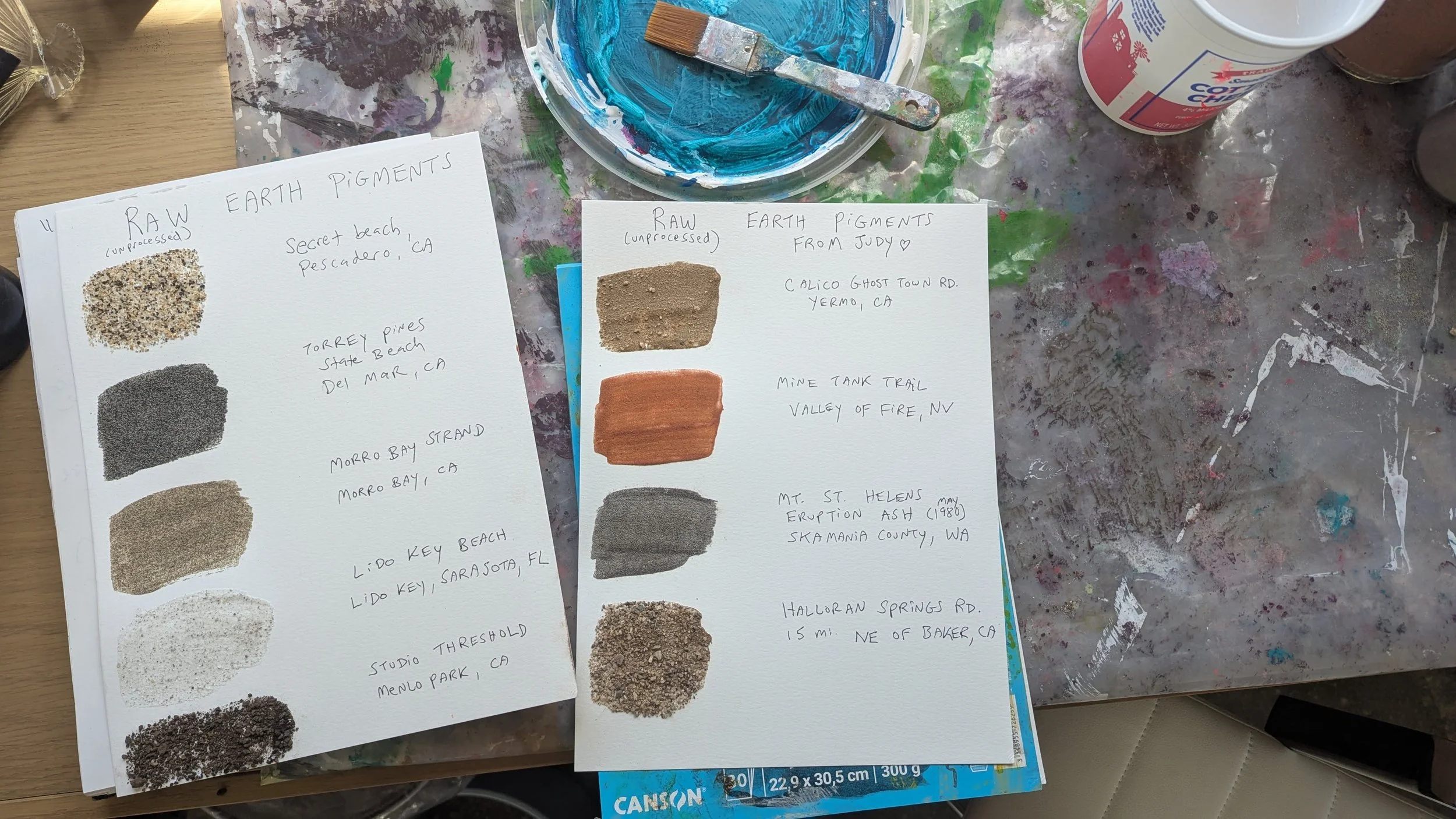 Two sheets of paper showing samples of raw earth pigments, each with handwritten labels and geographic locations. The left sheet lists unprocessed earth pigments from California beaches and human-made settings, with color swatches of sands and soils.