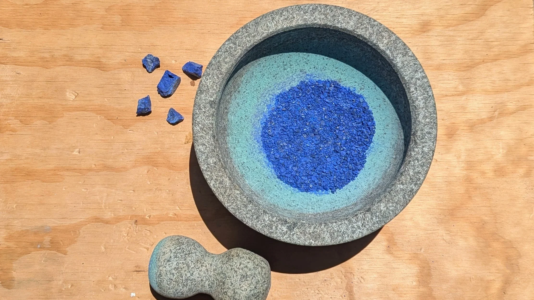 Gray stone bowl with blue granules inside, a few blue stones outside, and a small gray stone pestle on a wooden surface.