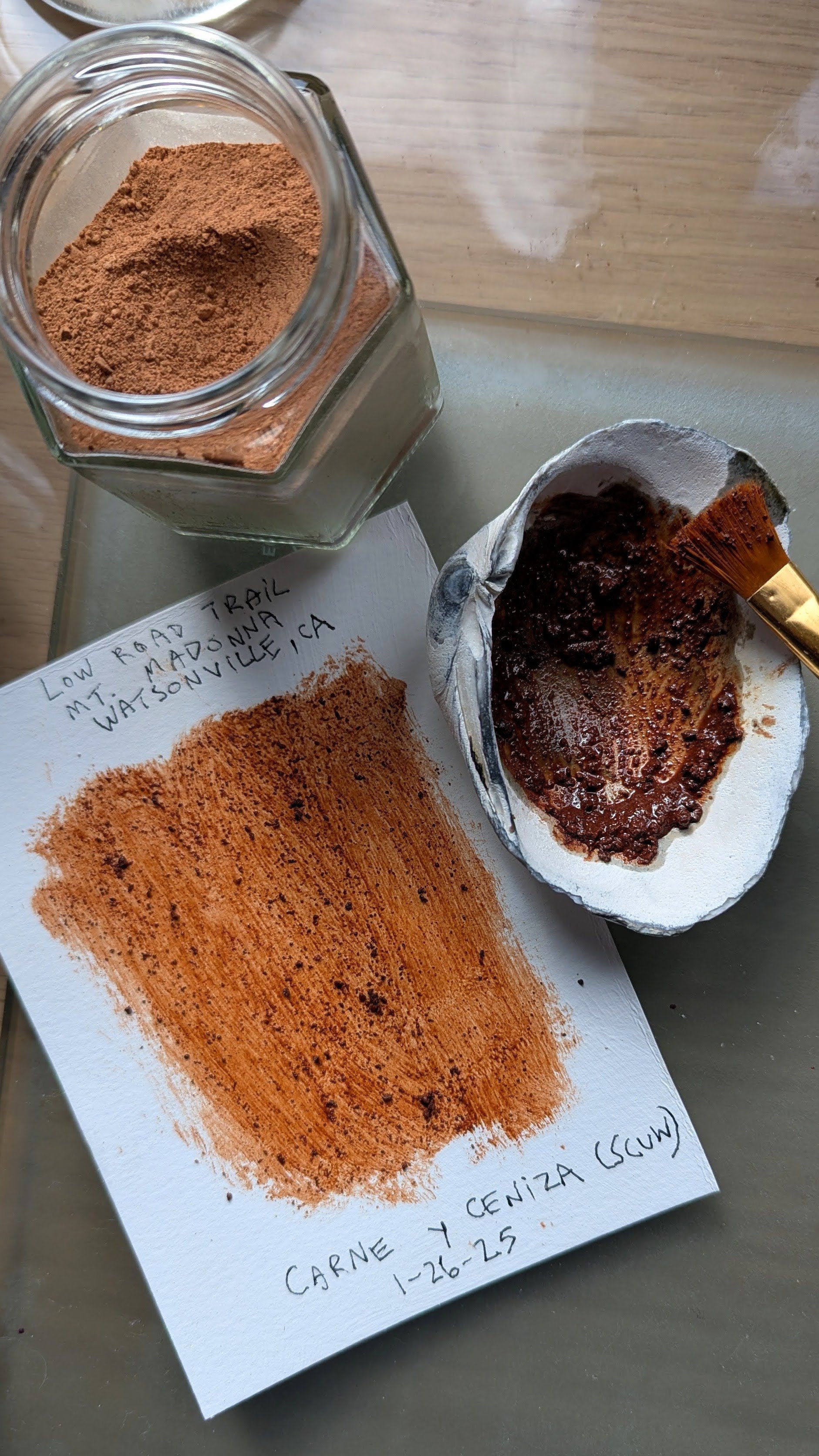 A jar of homemade earth pigment, a textured piece of paper with handwritten notes and a swatch of color, a small shell used as a paint palette, and a paintbrush resting on the shell.