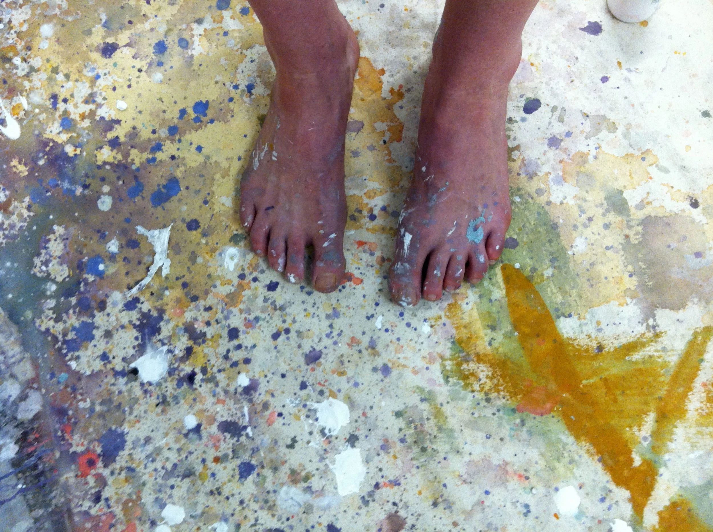 The artist's bare feet on a colorful studio floor splattered with paint stains.