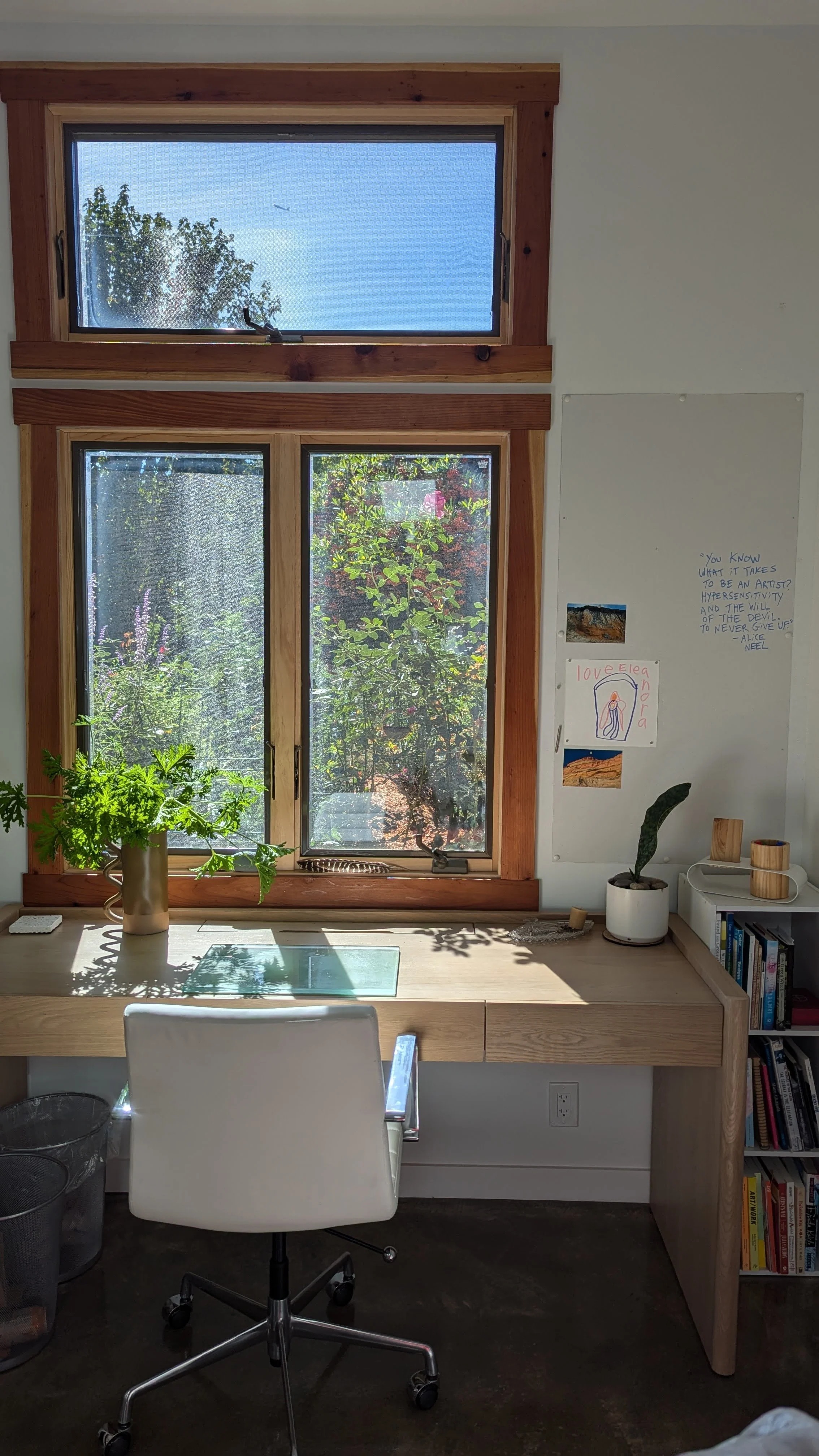 Artist's studio with a wooden desk and white swivel chair, large windows revealing greenery outside, potted plants on the desk, books on a small bookshelf, and artwork on the white wall.