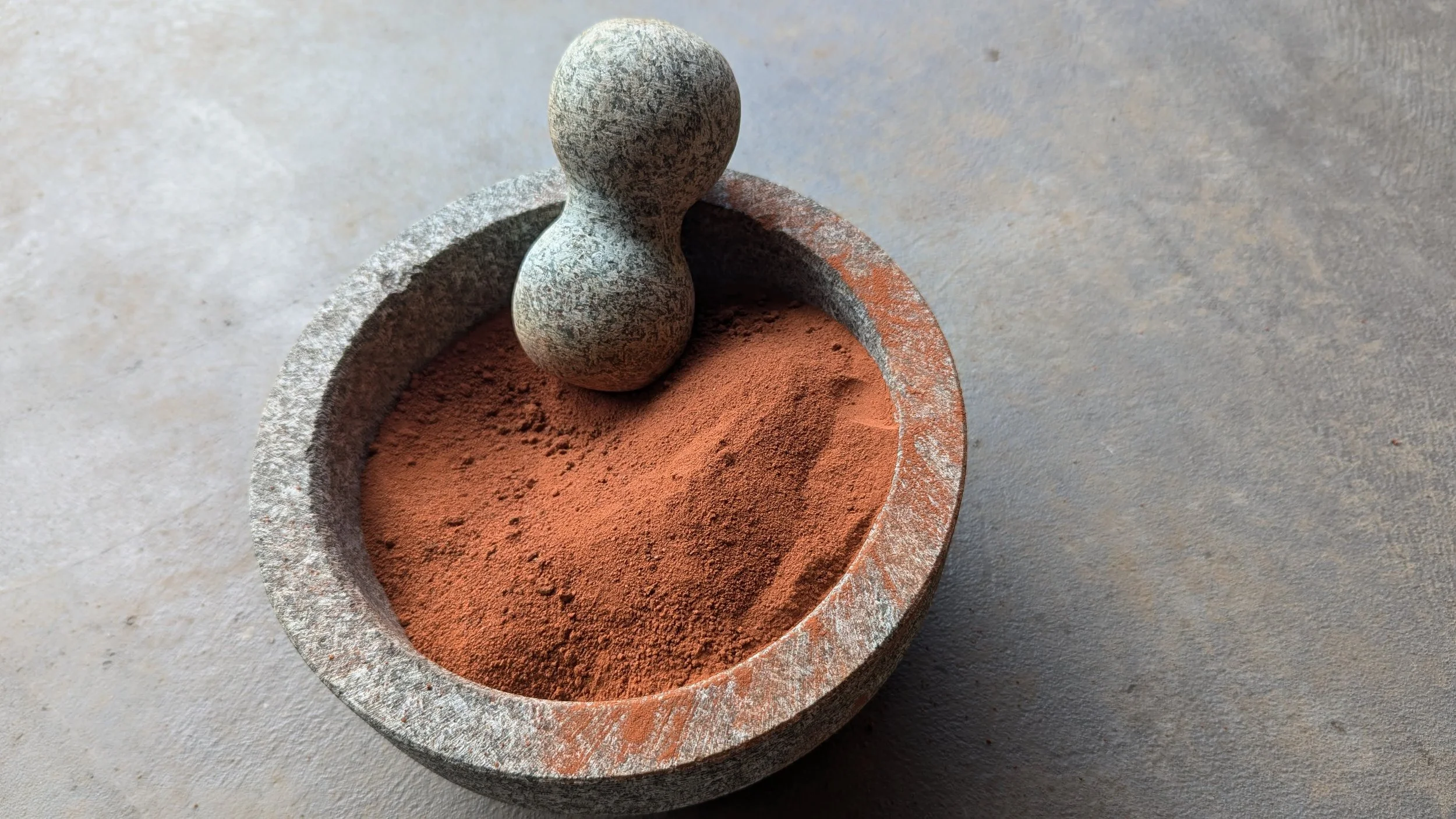 A stone mortar with a pestle, containing red powdered pigment on a neutral-colored surface.