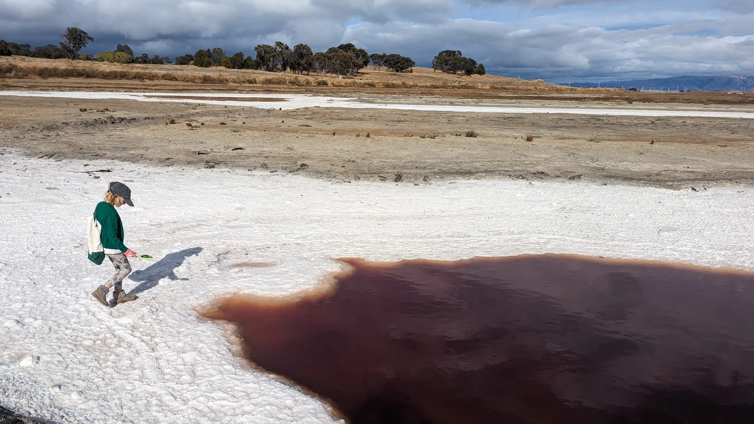 A child dressed in a green jacket, patterned leggings, and boots, wearing a hat and pink gloves, standing near a large pool of dark brown water on white salt flats with a landscape of brown hills and trees under a cloudy sky in the background.