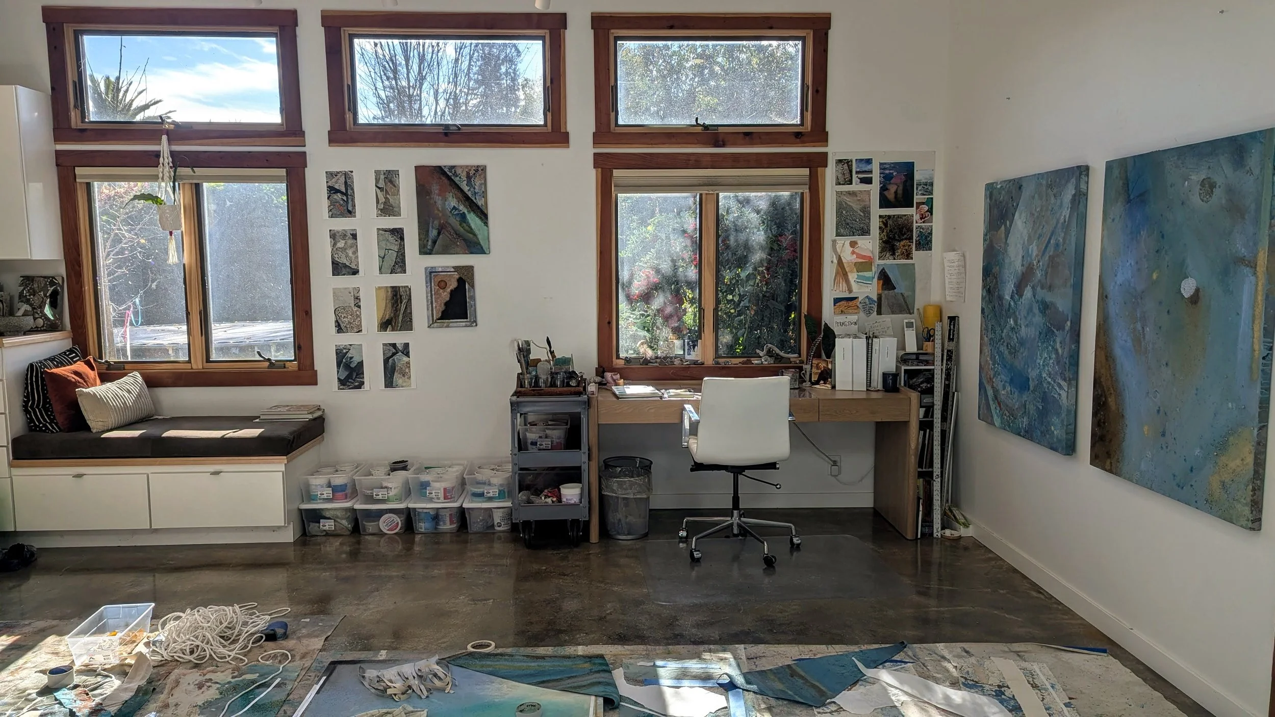 Art studio with large windows, featuring abstract paintings on the walls, a white chair at a desk, art supplies, and a cozy window seat with pillows.