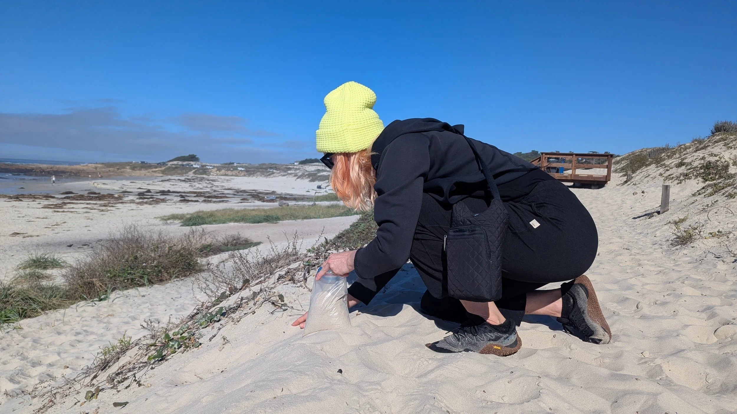 Artist Ahna Serendren wearing a neon yellow beanie, black jacket, and black pants crouching on sandy beach, using a clear plastic bag to collect sand, with dunes and blue sky in background.
