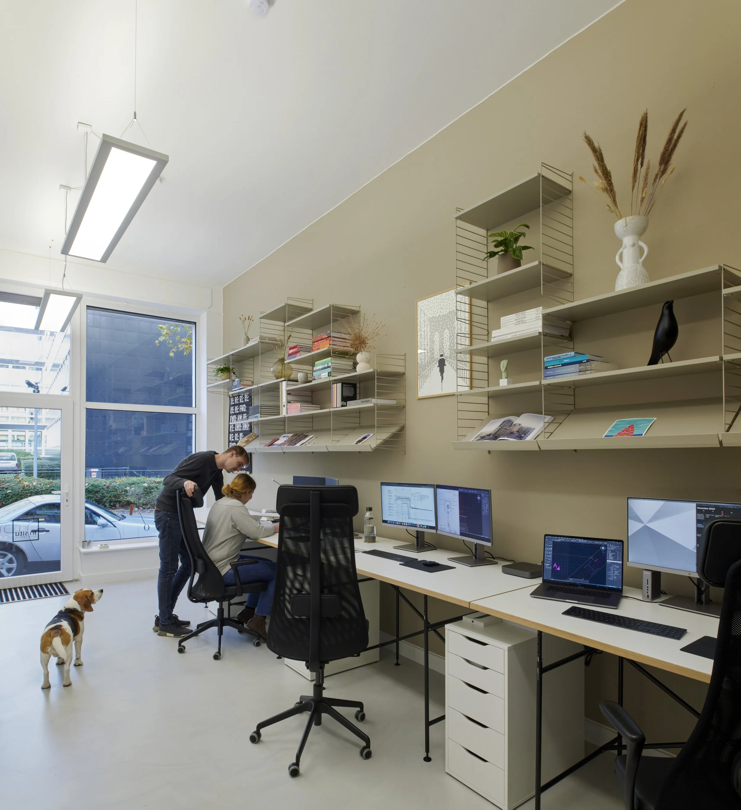 Ein modernes Büro mit mehreren Computern, Wandregalen mit Büchern und Dekorationsgegenständen, einem großen Fenster, einem Mann, der mit einer Frau am Schreibtisch spricht, und einem Hund im Raum.