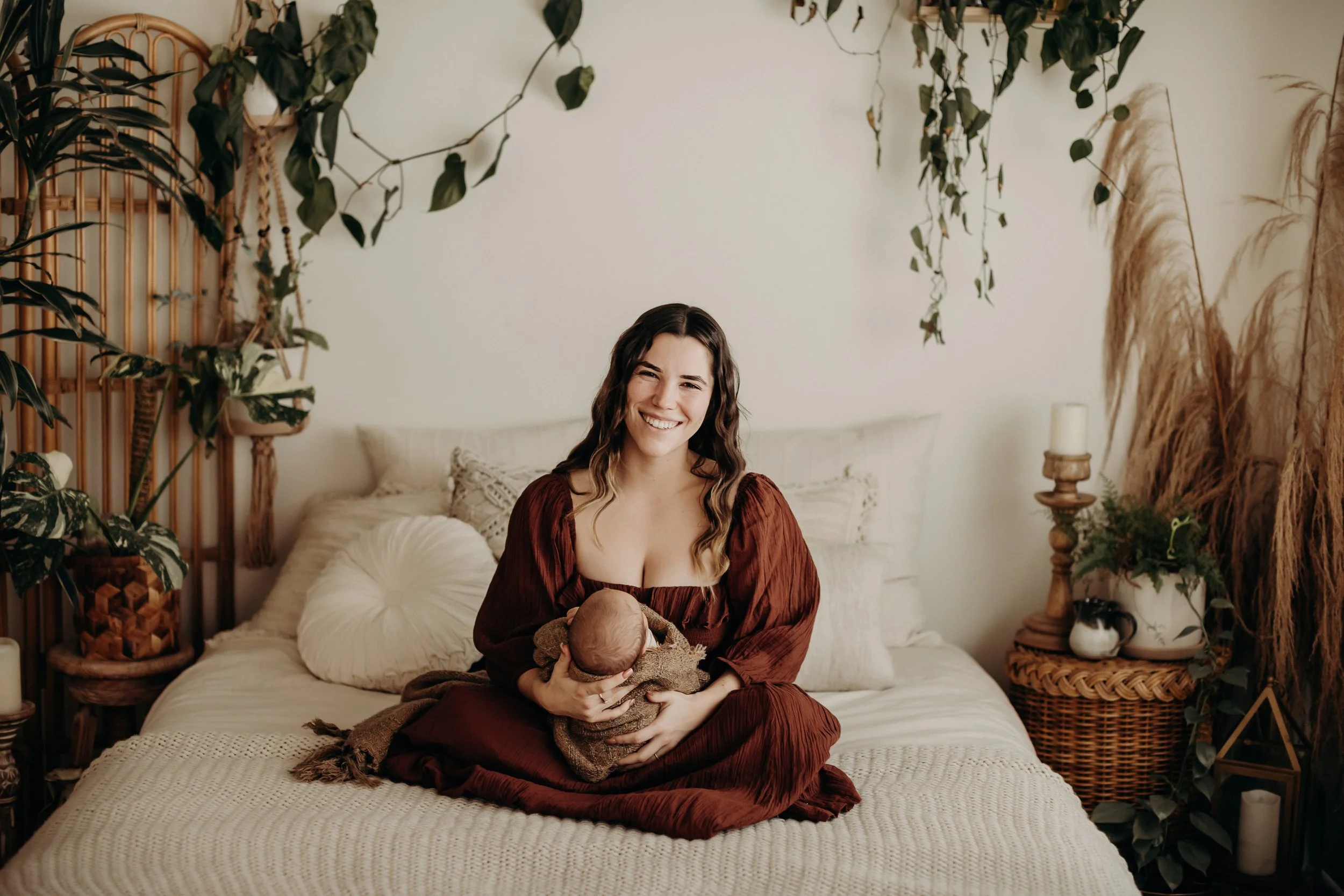 A woman sitting on a bed holding a newborn baby, smiling at the camera, in a cozy room decorated with plants and candles.