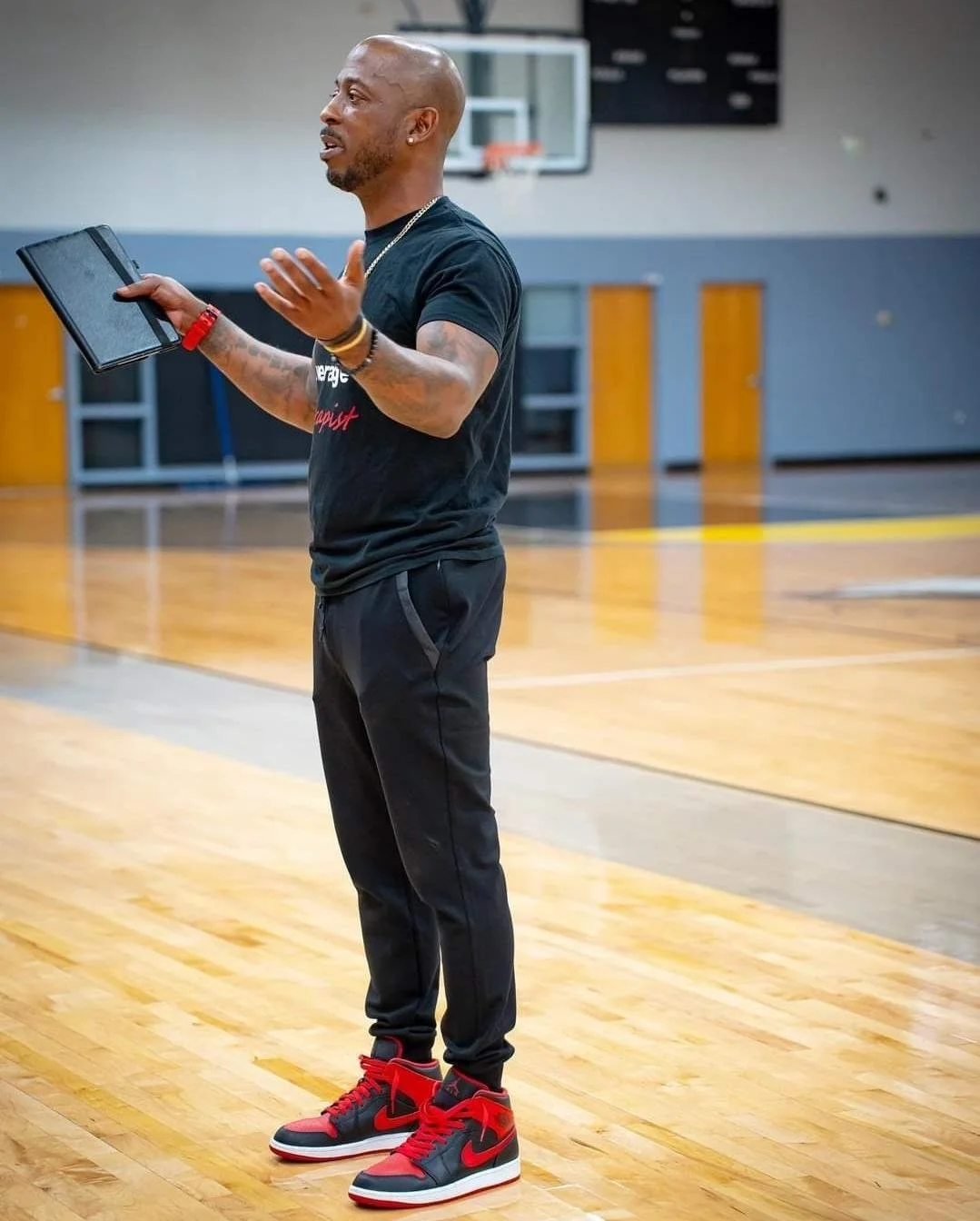 A man standing on a basketball court, holding a tablet, and gesturing while speaking. He is wearing black athletic clothes and red and black sneakers in a gymnasium.