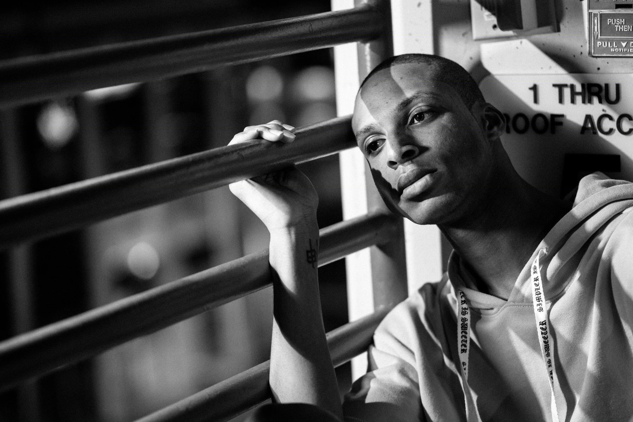 A young man with a shaved head sitting near a metal railing, resting his head on his hand as he stares thoughtfully into the distance. The photo is in black and white.