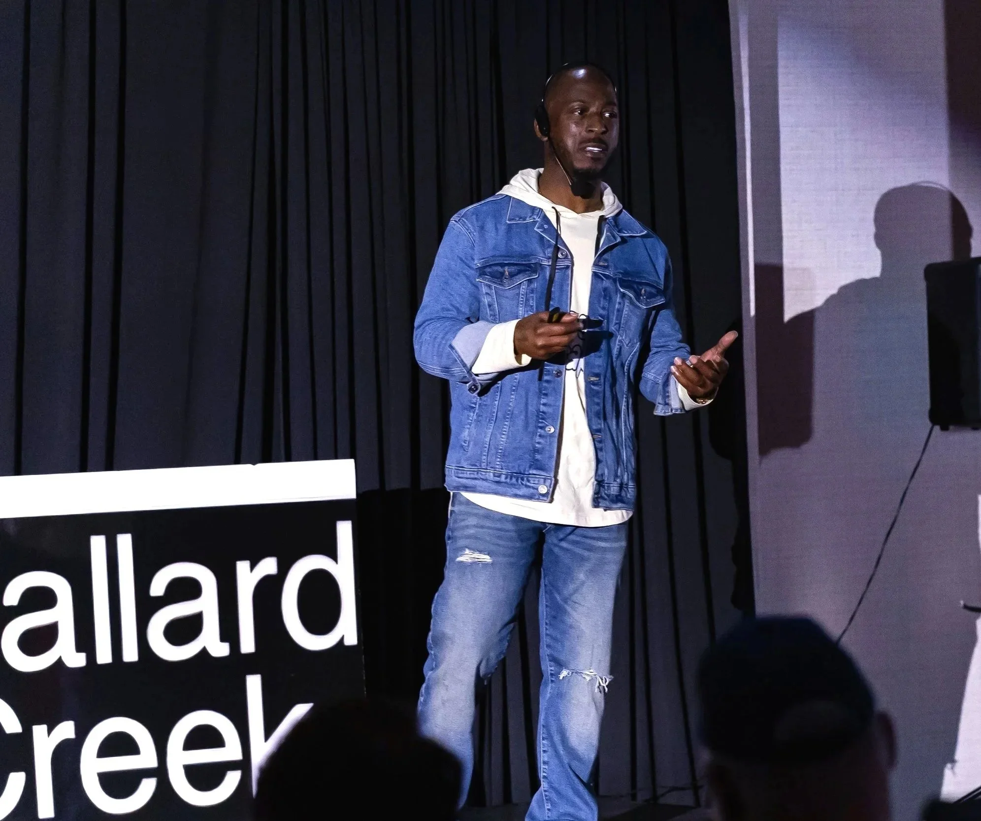 A man wearing a denim jacket and distressed jeans speaking on stage with a microphone and headset, standing next to a sign that reads 'Allard Creek,' with black curtains in the background.