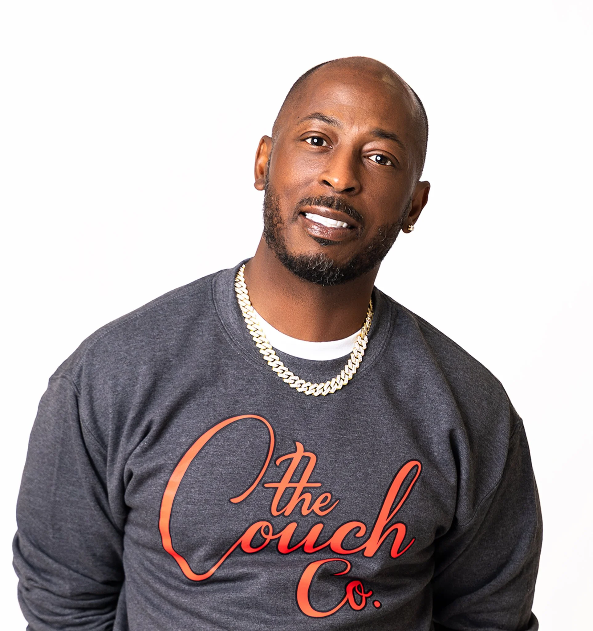 A man with a goatee and earrings, wearing a dark gray sweatshirt with red cursive text that reads 'The Crouch Co.', a gold chain, and a white shirt underneath, posing against a plain white background.
