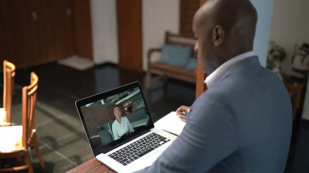 A man in a gray suit participating in a video call on a laptop in a home office.