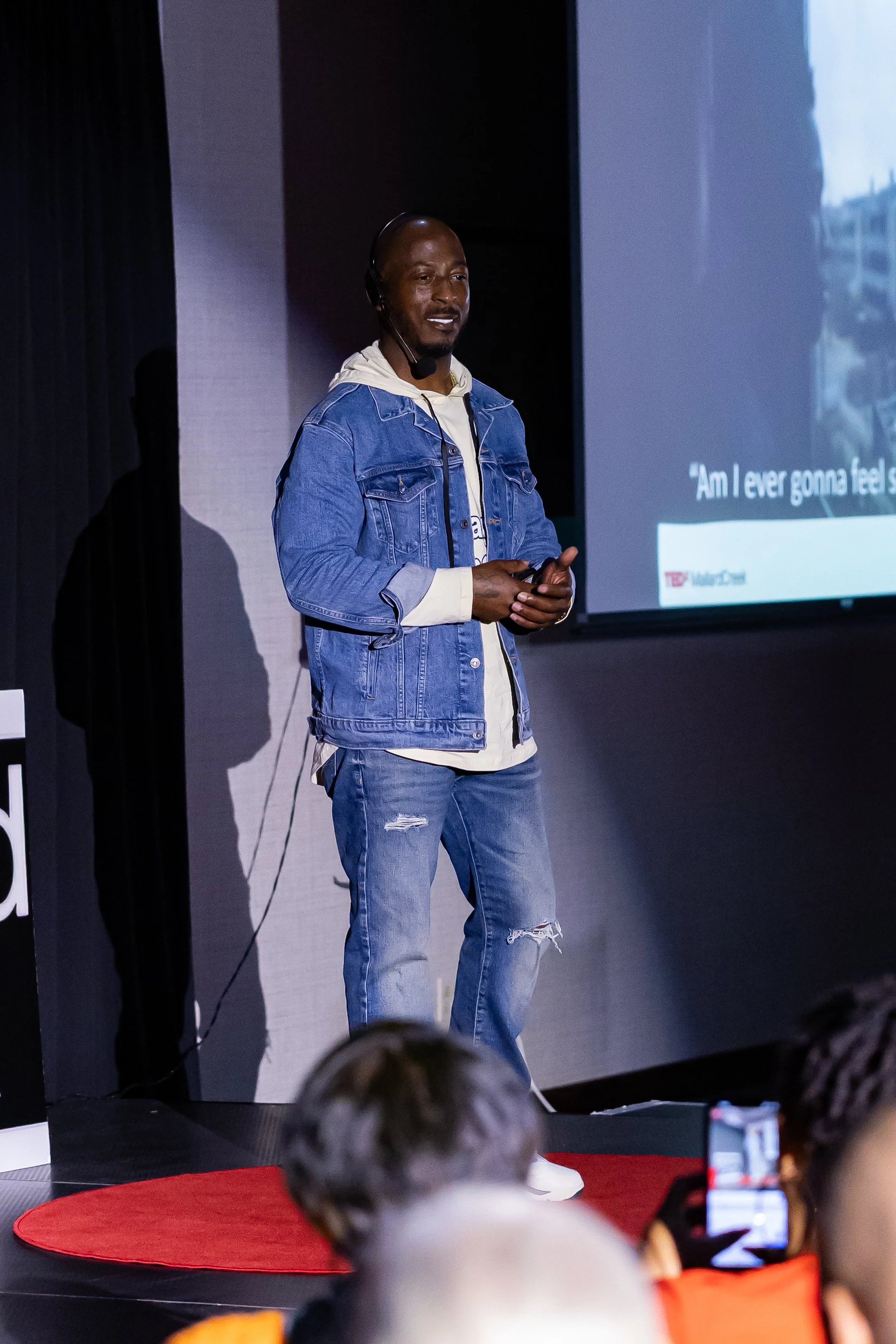 Man wearing a denim jacket and distressed jeans giving a presentation on stage with a large screen behind him and audience members in the foreground.