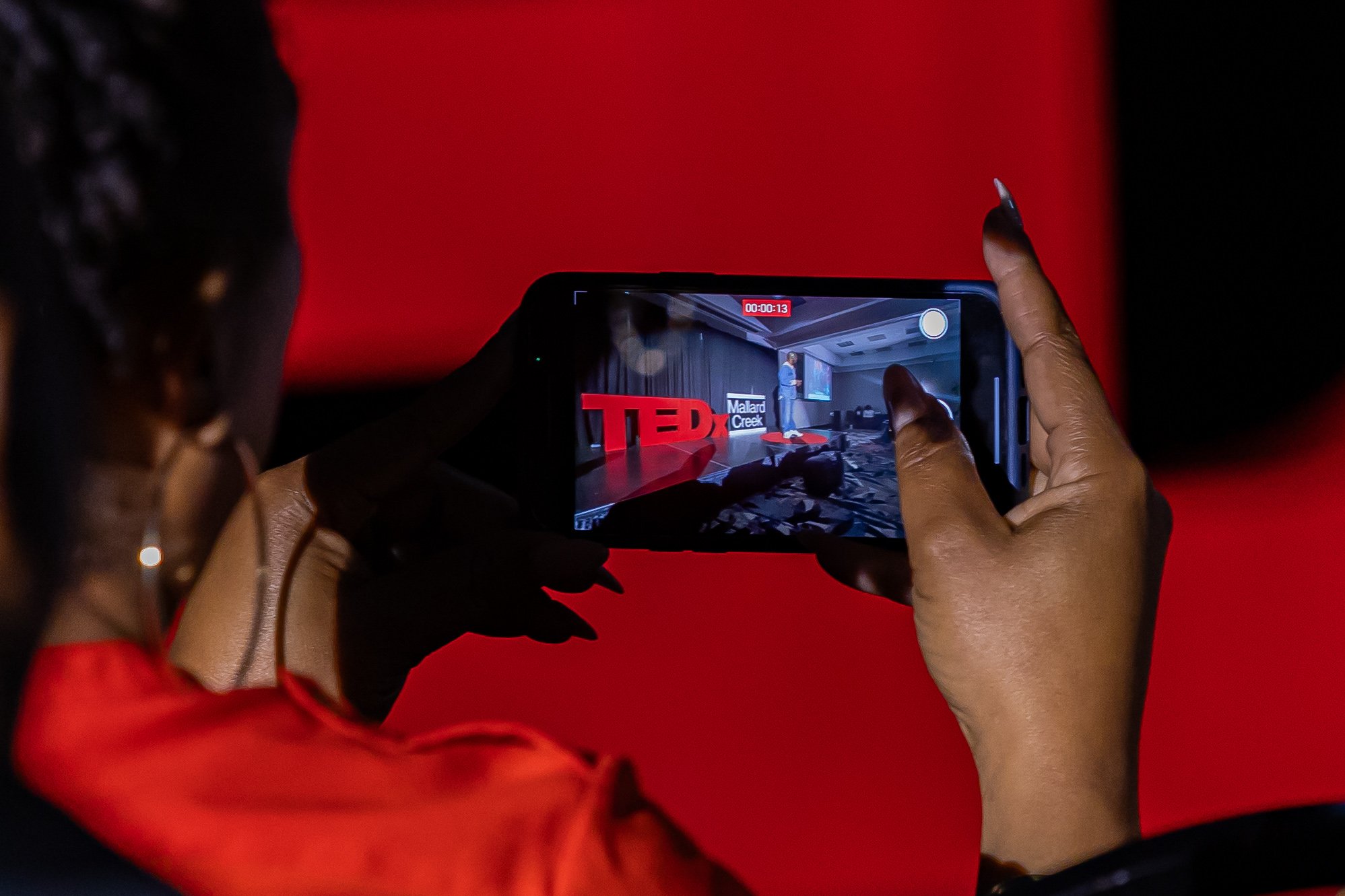 Person recording a TEDx event presentation on a smartphone in a dimly lit room with a red background.