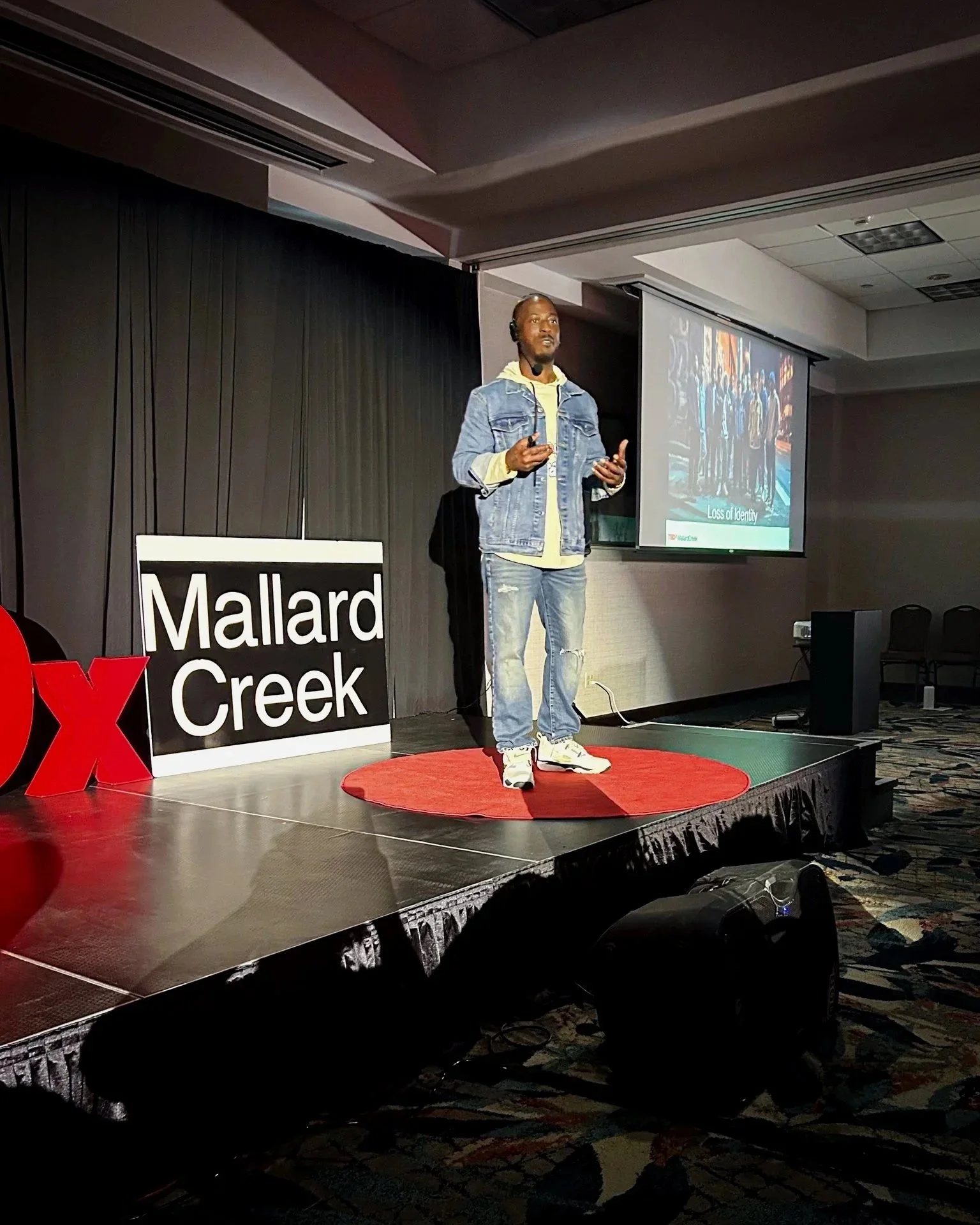 A man giving a presentation on stage at Mallard Creek, with a screen showing slides behind him, and a large 'O' and 'X' sign nearby.