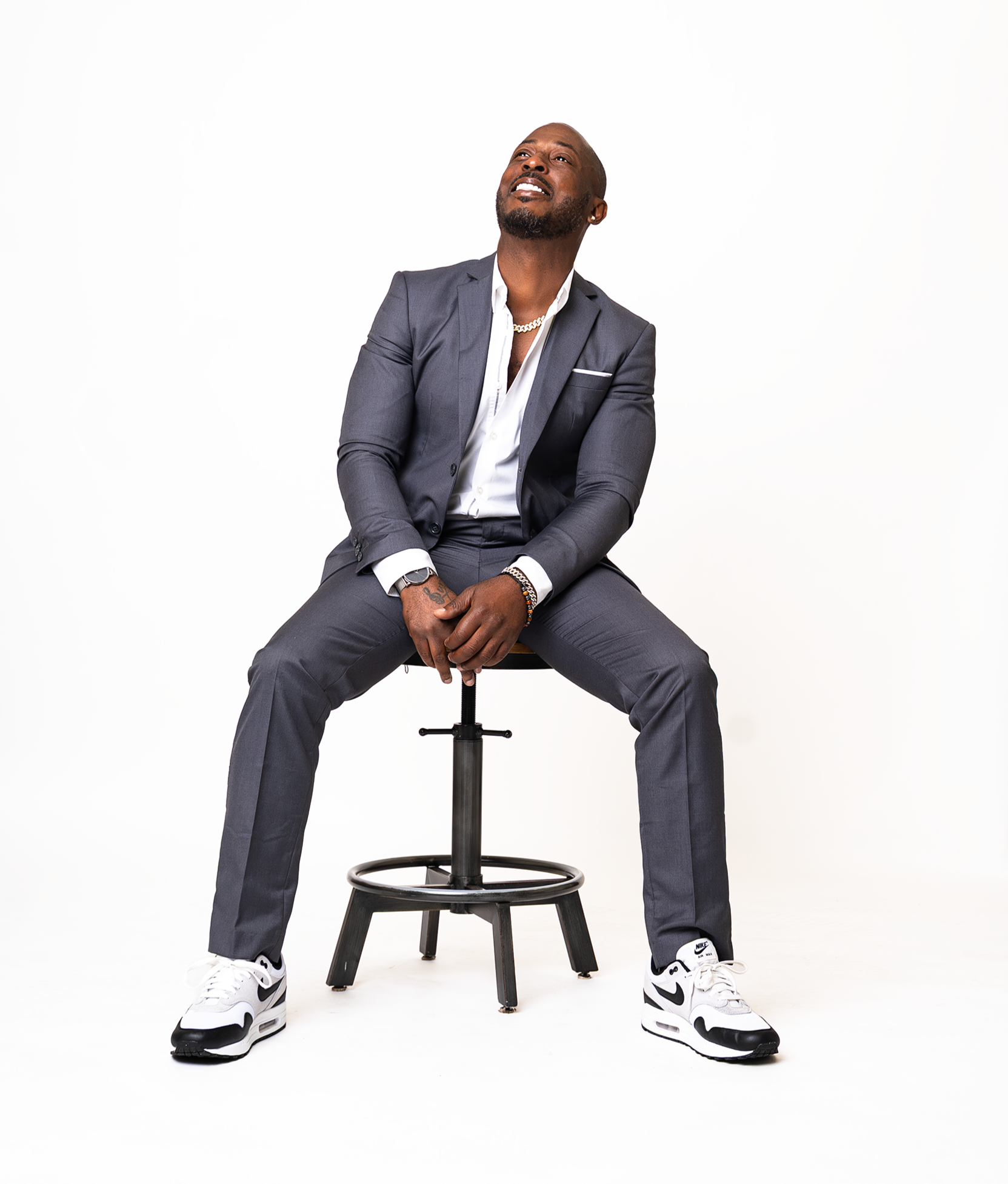 Man in a gray suit sitting on a stool with legs apart, looking upwards, wearing white and black Nike sneakers, with a white shirt, gold chain, and bracelets, against a plain white background.