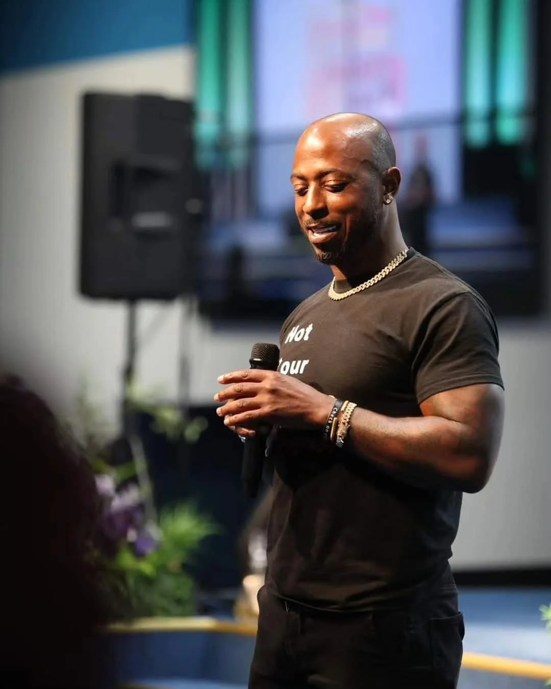 A man with a shaved head wearing a black T-shirt and jewelry, holding a microphone, standing in front of a large screen and speaker in an indoor setting.