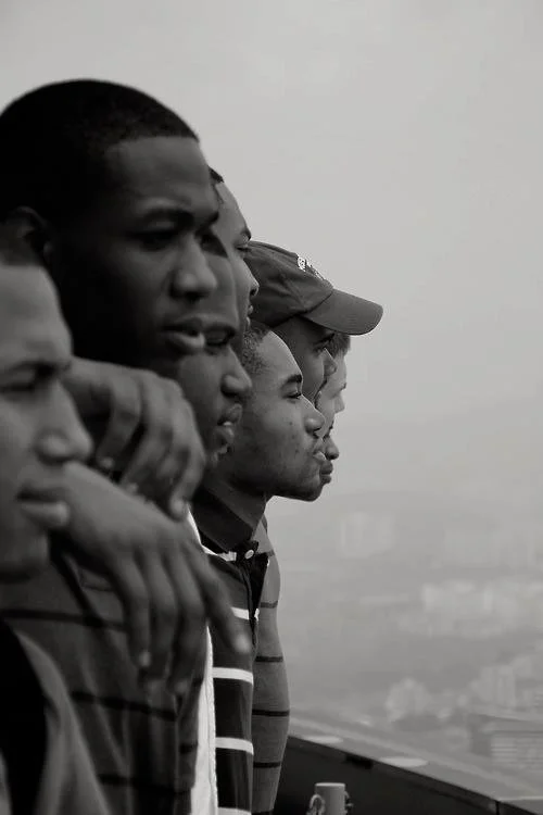 A black and white photo of five young men standing in a row, looking to the right, with a cityscape in the background.