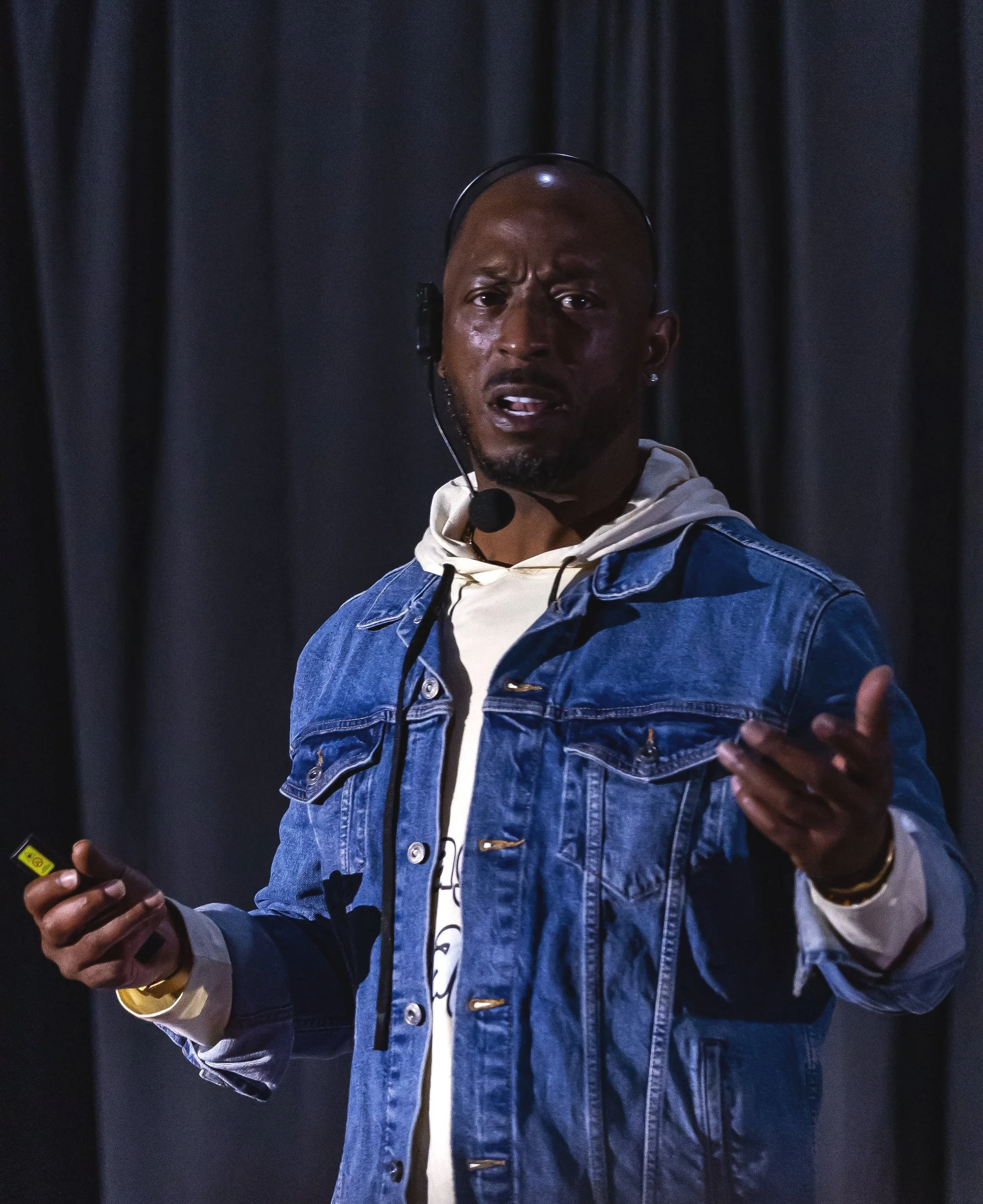 A man speaking on stage, wearing a denim jacket, hoodie, and a headset microphone, with a dark curtain background.