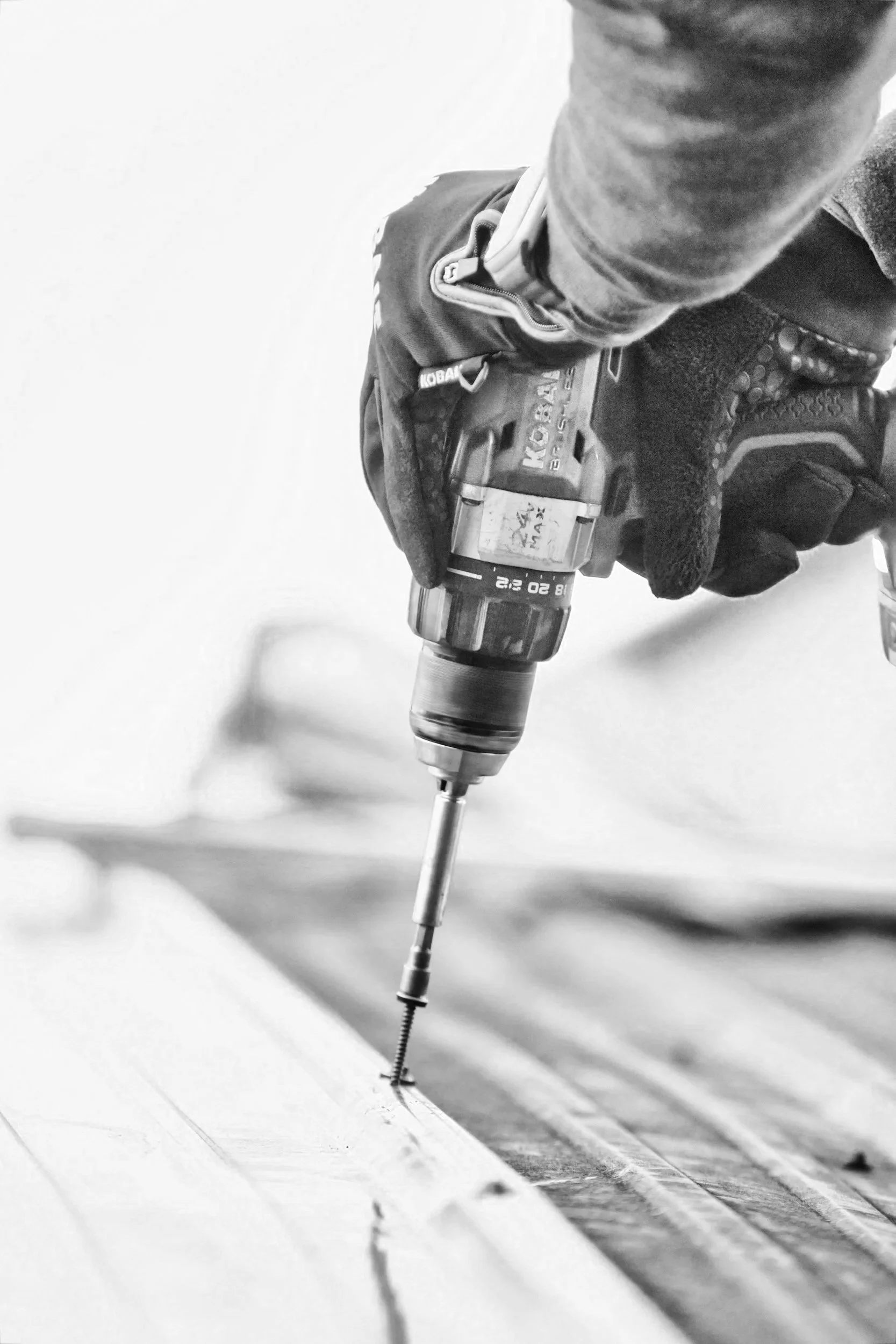 Close-up of a person using a cordless drill to drive a screw into a piece of wood. The person is wearing gloves and a long-sleeve shirt, and the image is in black and white.