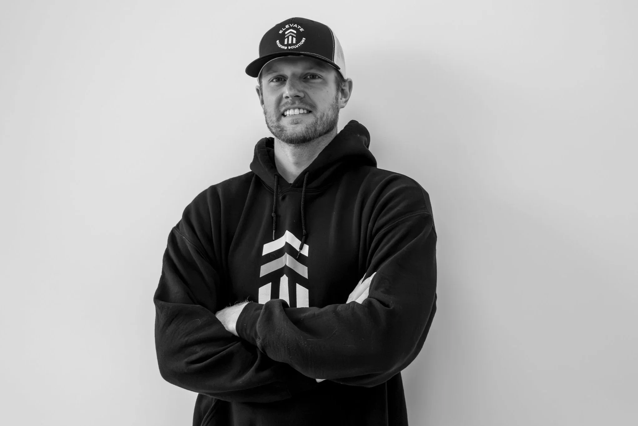 A man wearing a cap and hoodie with crossed arms, standing against a plain wall.