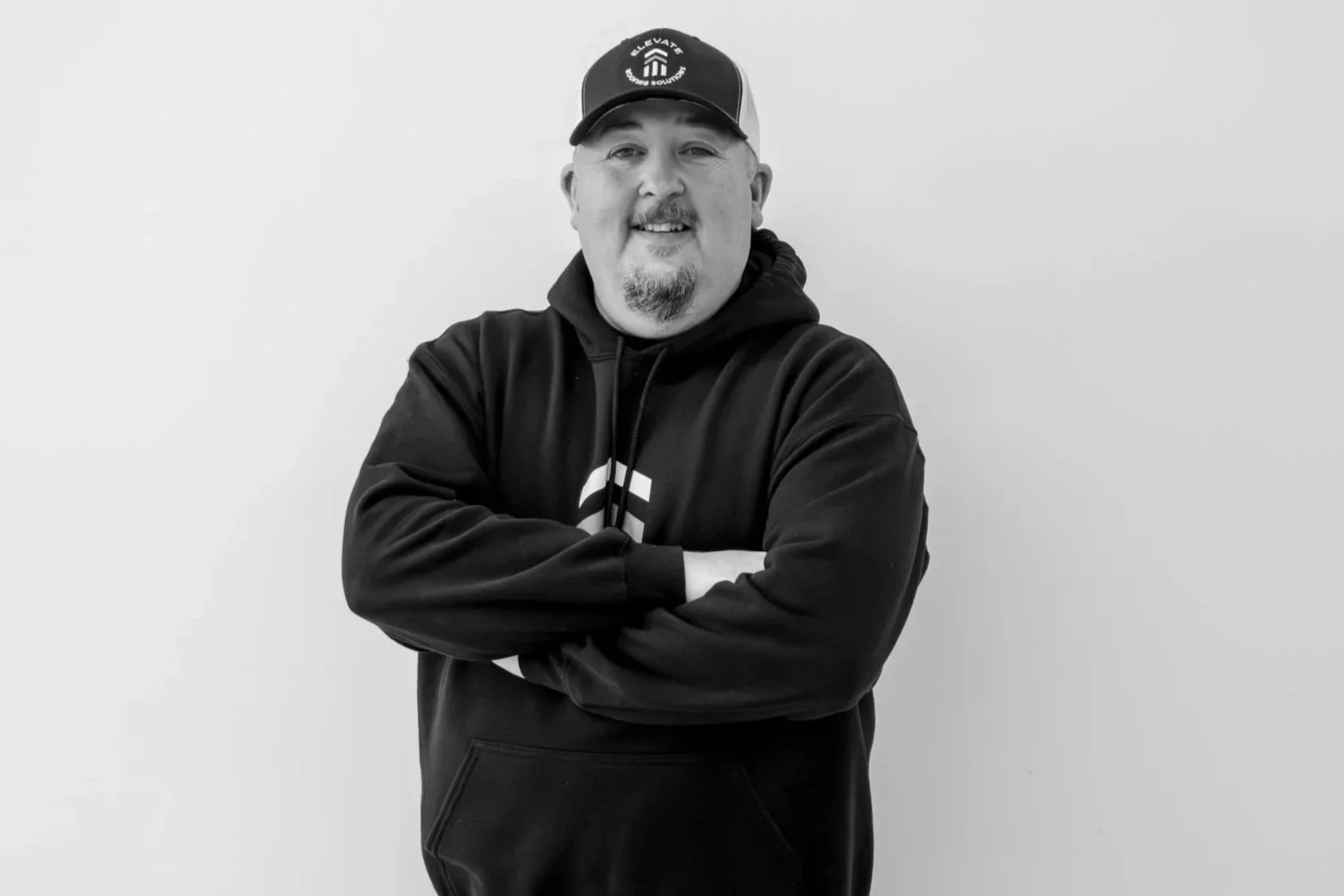 A man with a beard and mustache wearing a baseball cap and hoodie, crossing his arms and smiling against a plain wall.