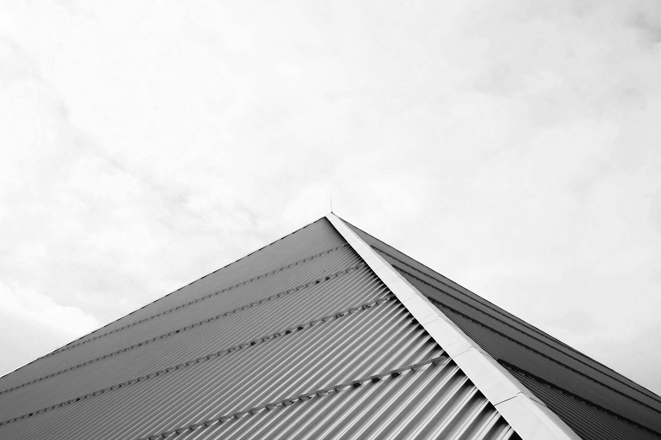 Black and white photo of the corner of a metallic building with a pointed roof, sky in the background.