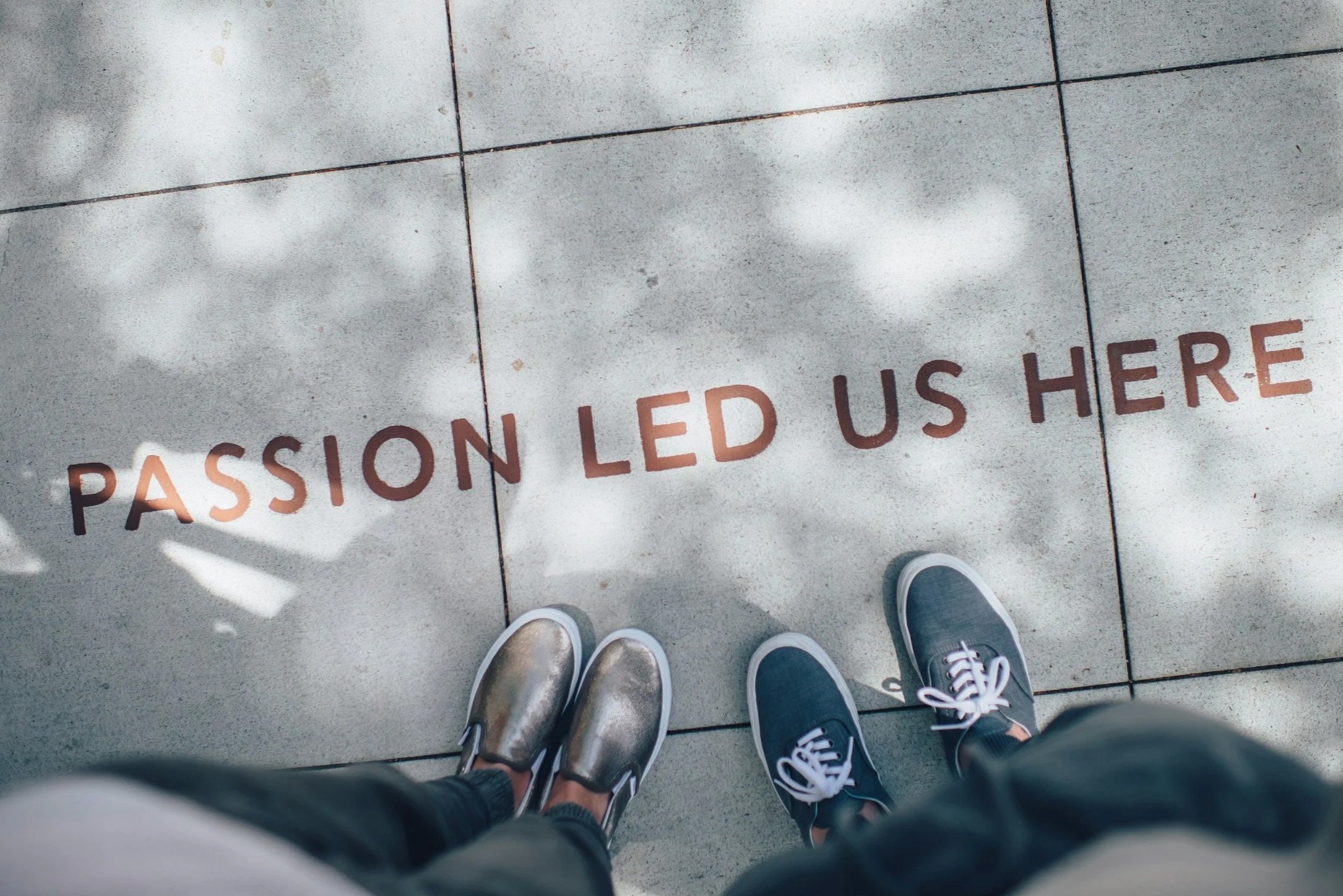 Two people standing on a concrete sidewalk with the words "PASSION LED US HERE" written on the ground in brown letters, with shadows of leaves overhead.