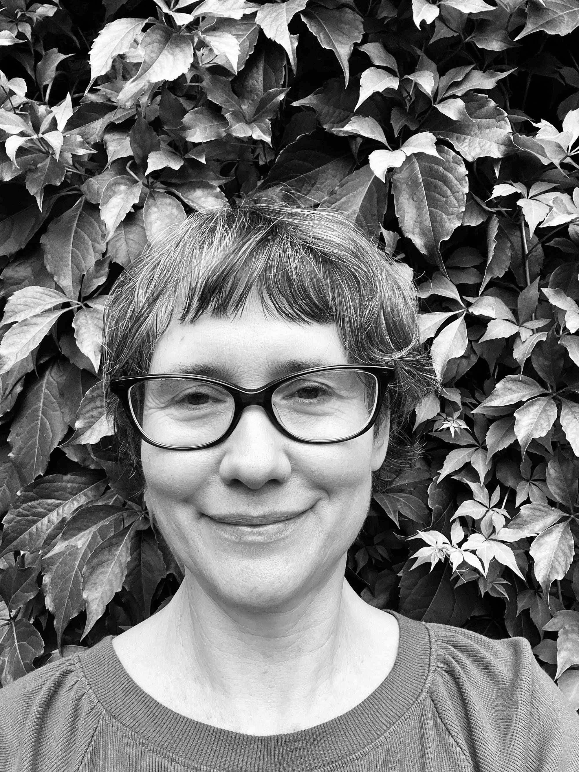 A woman with short hair and glasses, smiling, stands in front of a wall of leafy plants, in a black-and-white photograph.