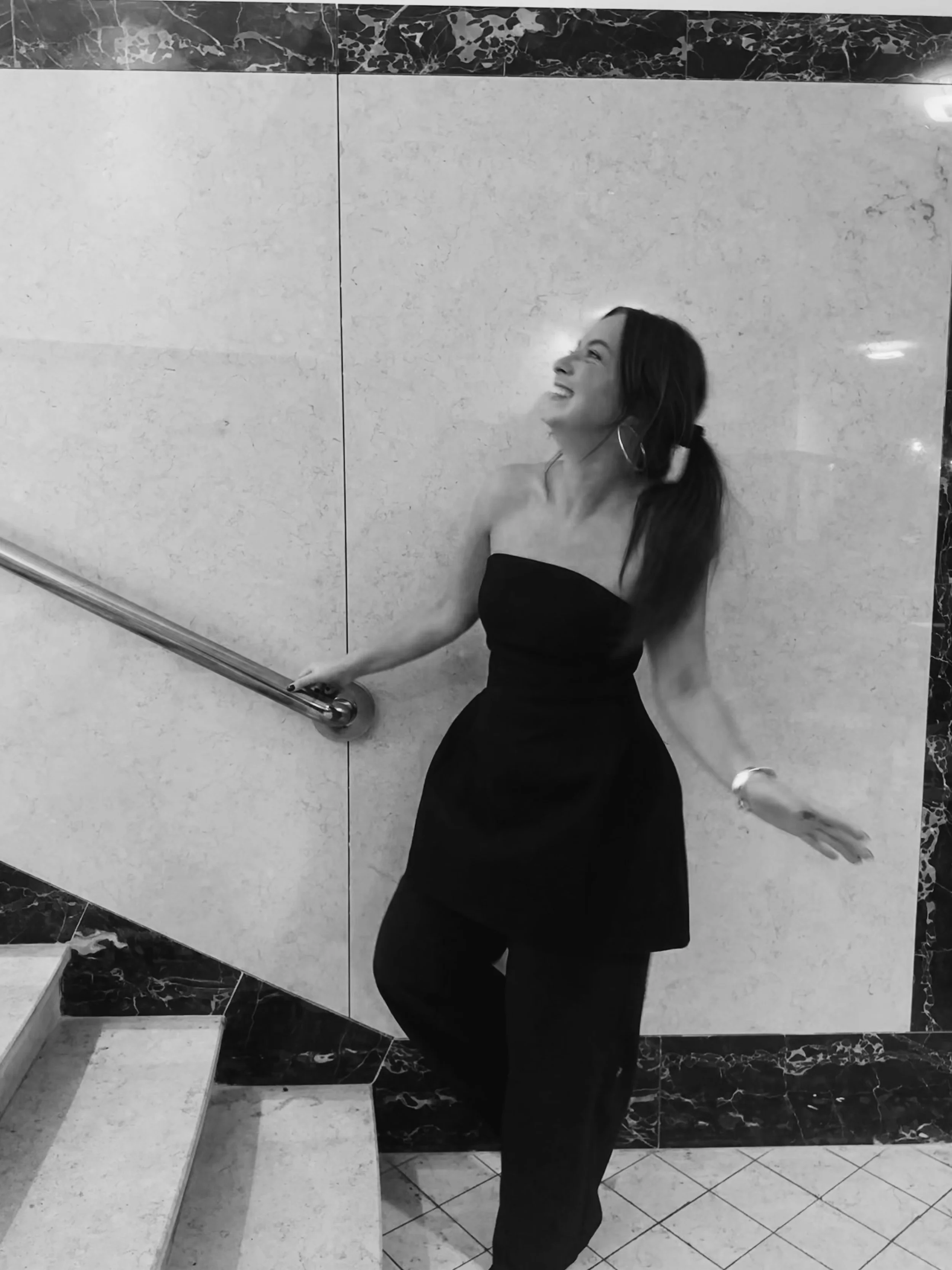 A woman in a black dress and pants laughing and posing against a tiled wall, holding a metal handrail.