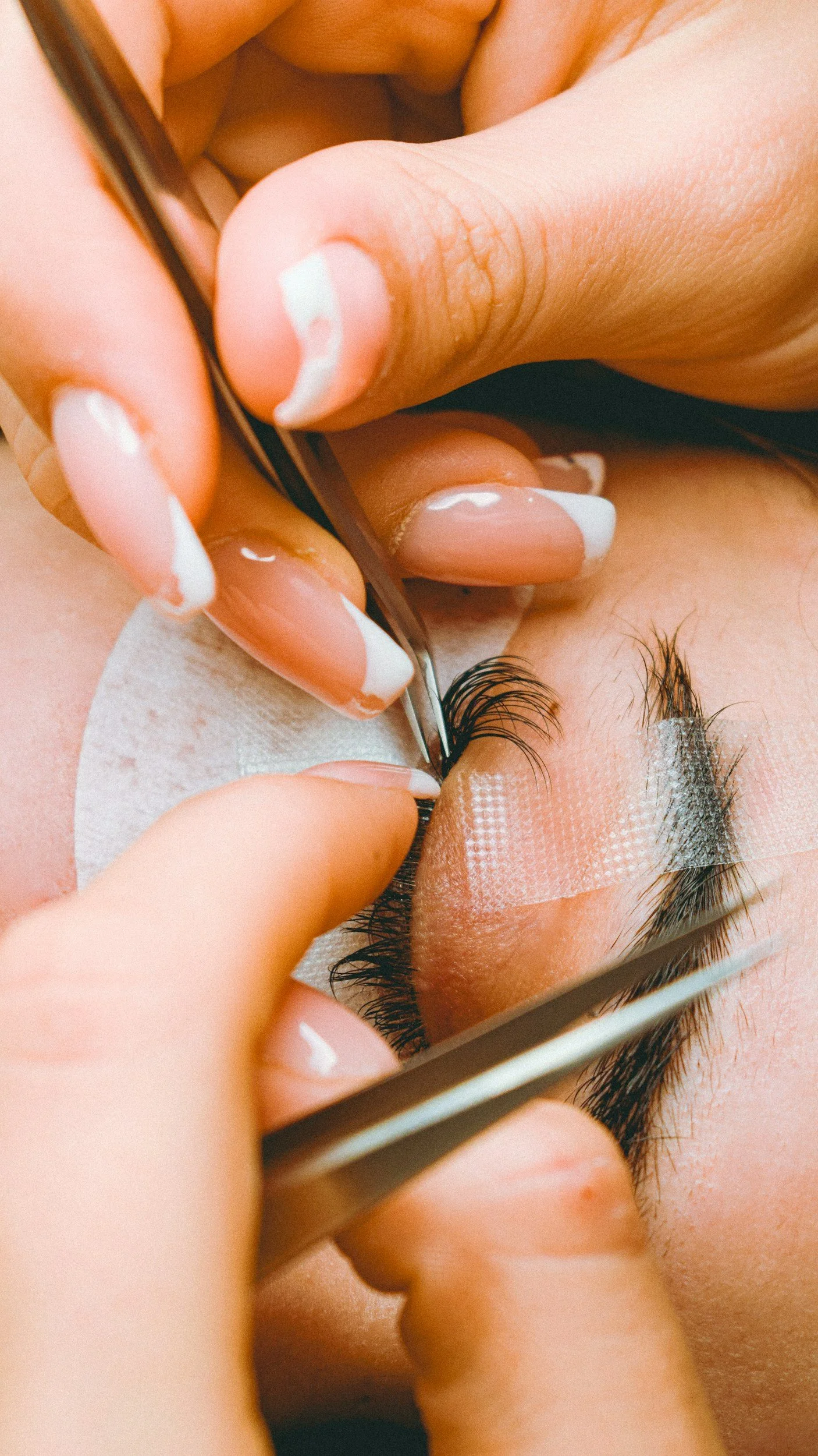 Close-up of a person receiving eyelash extension application, with technician using tweezers to attach individual lashes to natural eyelashes, using adhesive tape to hold eyelids open.