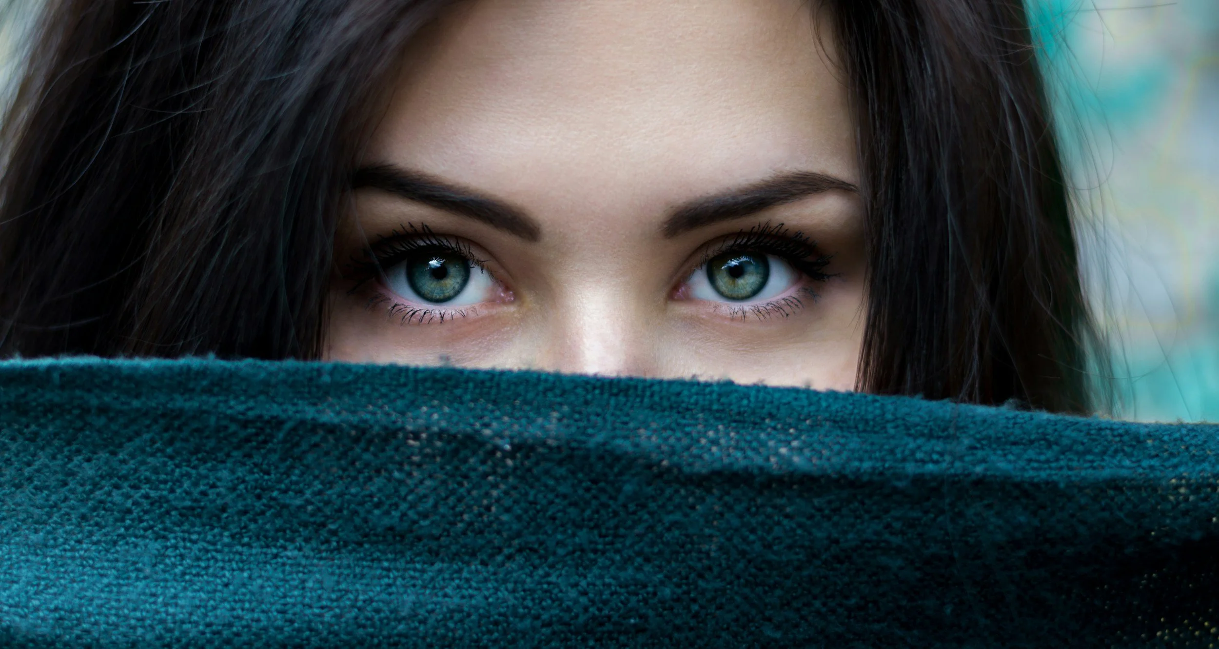Close-up of a woman with striking blue eyes and dark hair, partially covered by a teal fabric masking her lower face.