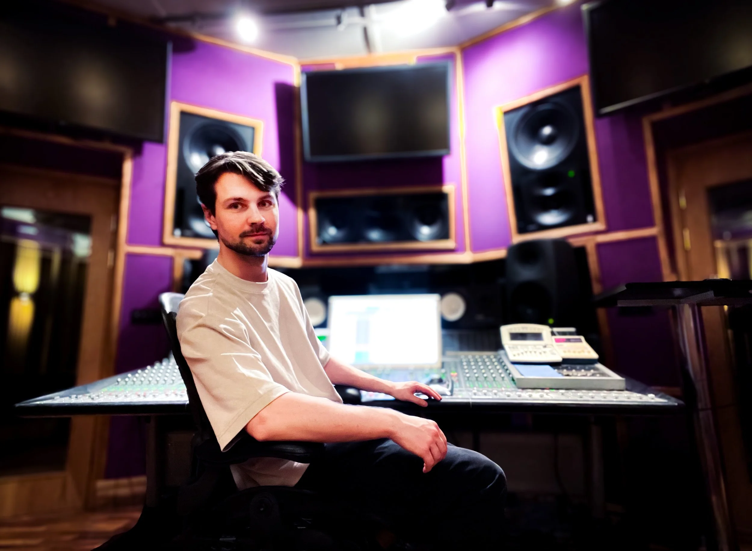 A man sitting in a recording studio surrounded by sound mixing equipment and large speakers, with purple acoustic panels on the walls.