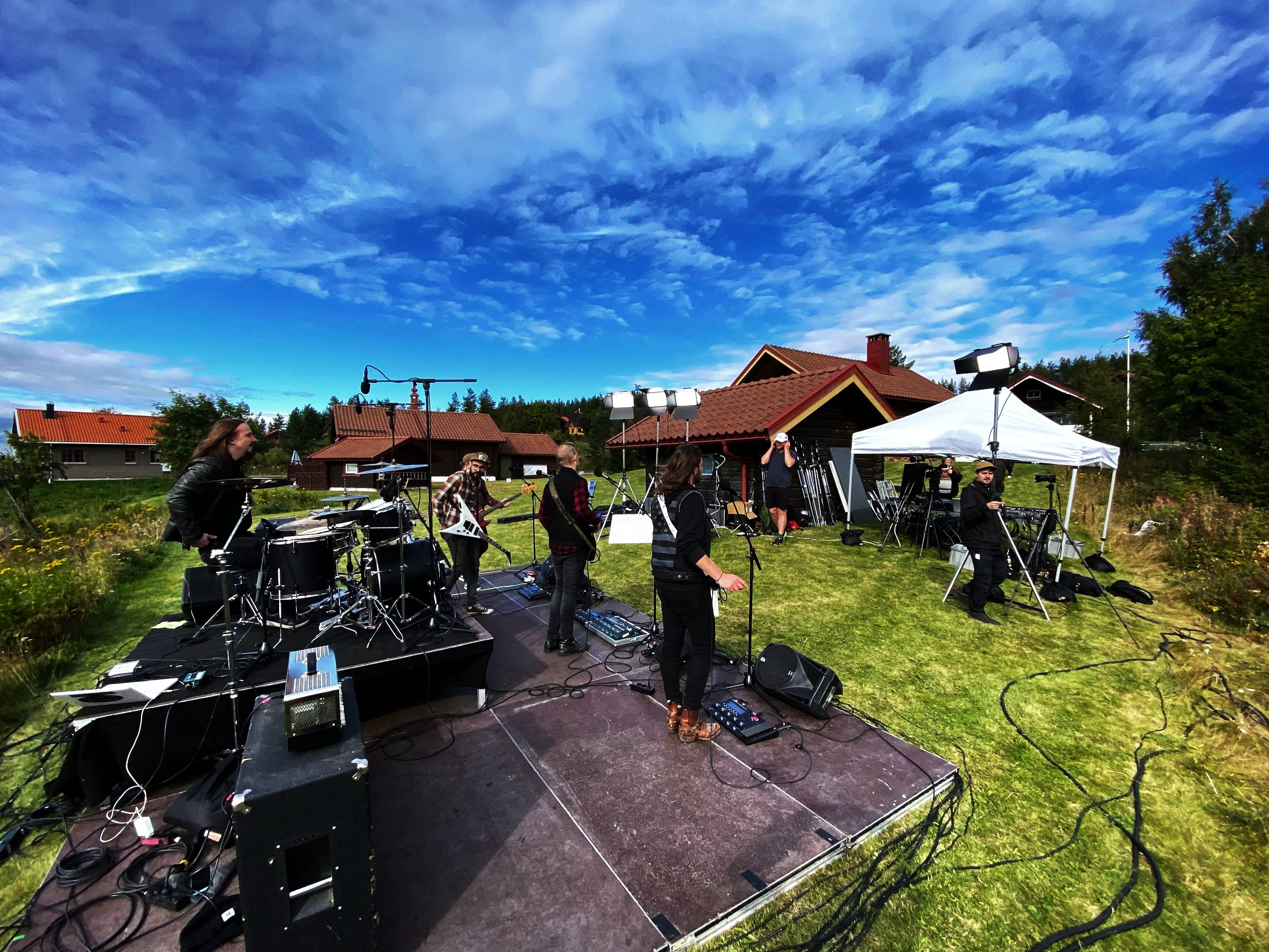 A band performs outdoors on a grassy hill under a partly cloudy blue sky, with houses and trees in the background. The band has multiple members playing various instruments, including drums, guitar, keyboards, and vocals. A white canopy covers some equipment, and there are stage lights and cables visible.