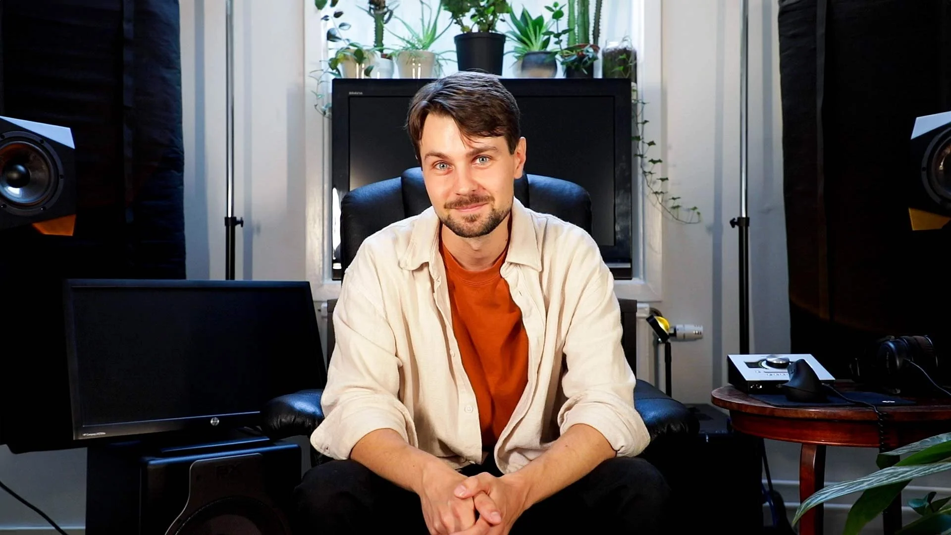 A man sitting in a recording studio with audio equipment, plants, and a television behind him.