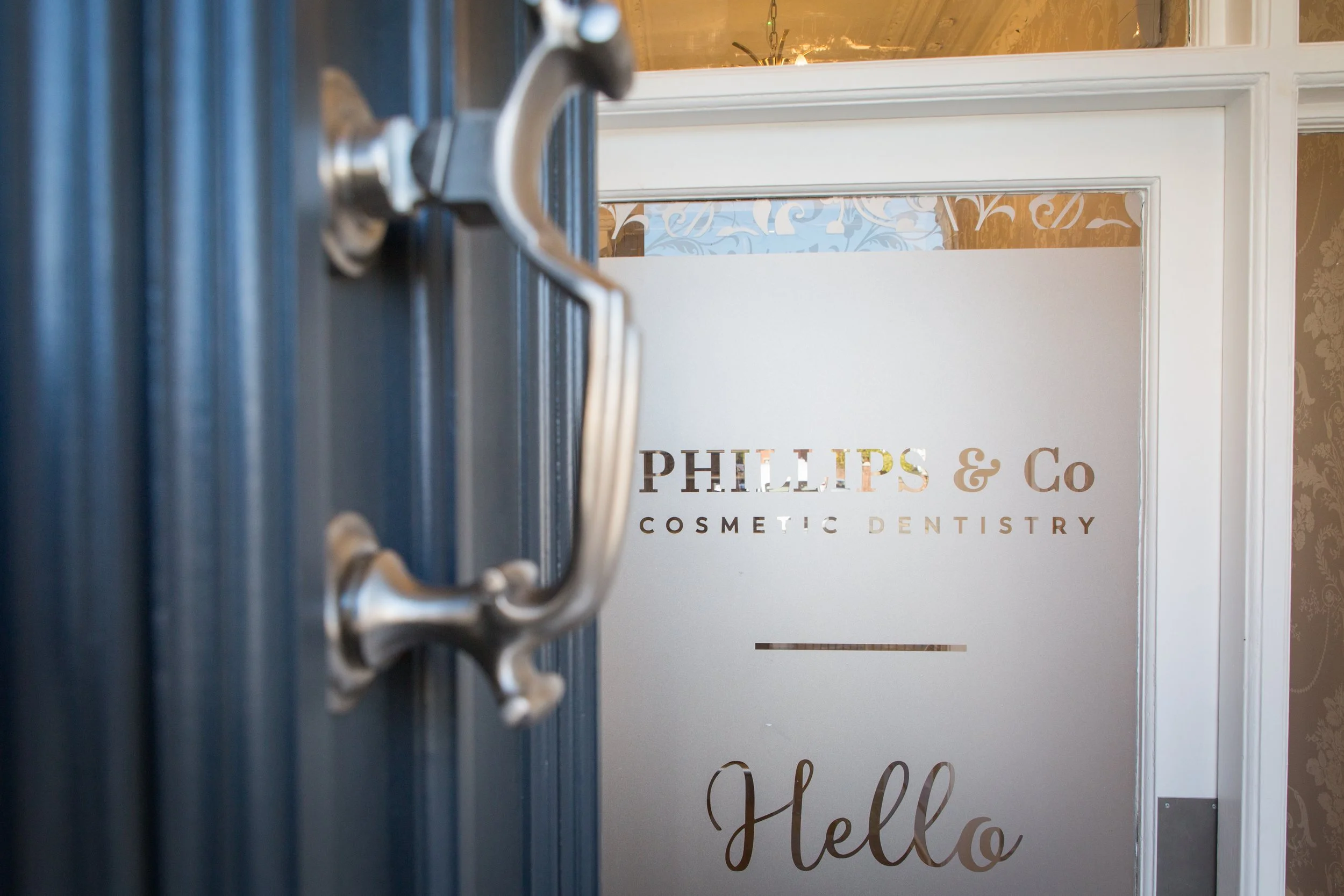 View of the entrance to Phillips & Co Cosmetic Dentistry with a frosted glass door that has the company's name and logo on it, and a blue door handle on the left.