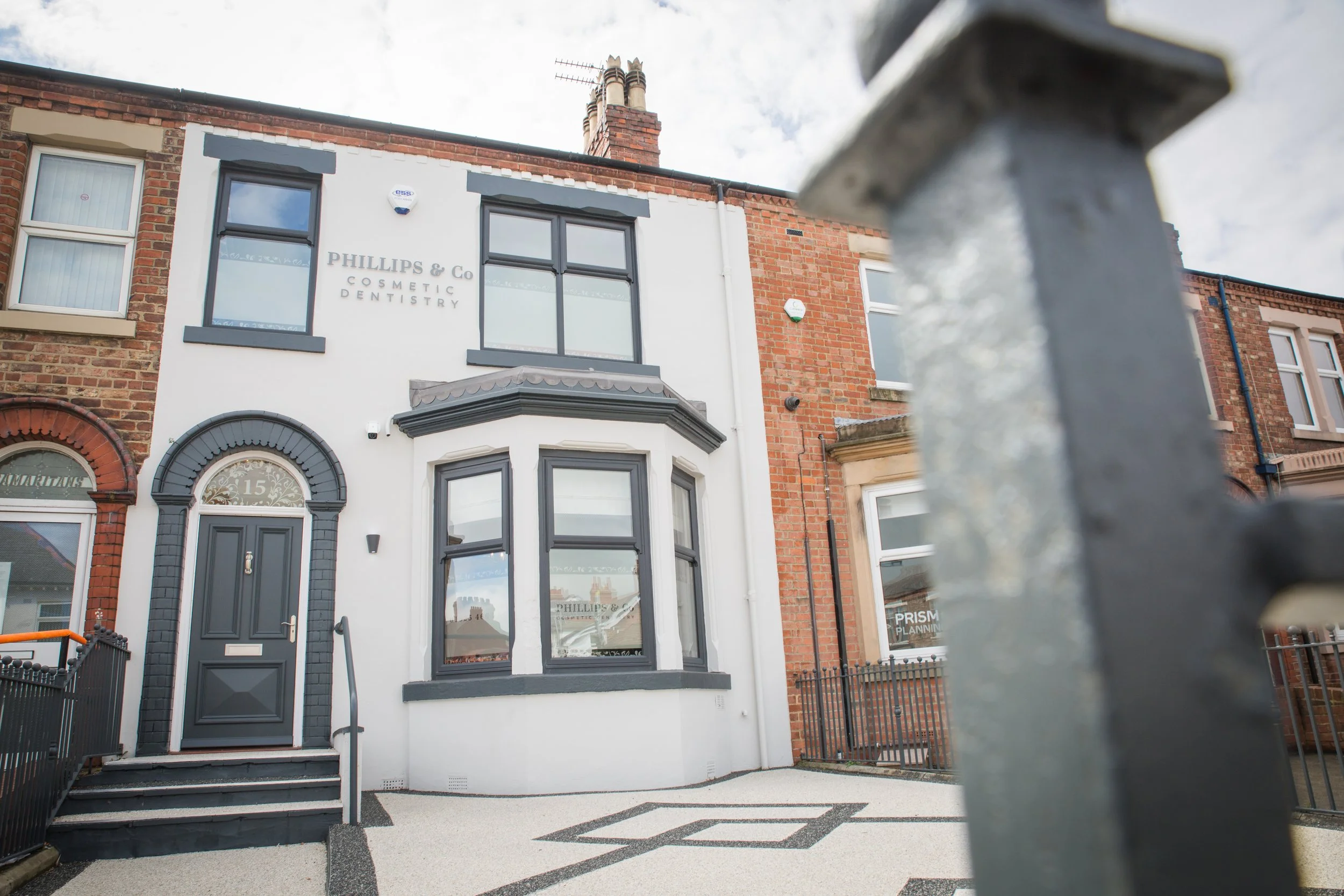 A white and gray building with a sign that reads 'PHILLIPS & Co COSMETIC DENTISTRY' on the front, located at 15. The building has large windows and a dark-colored front door with the house number on it. There is a small front yard with geometric design painted on the ground and a low black fence. Another building with a brick facade is adjacent on the right.