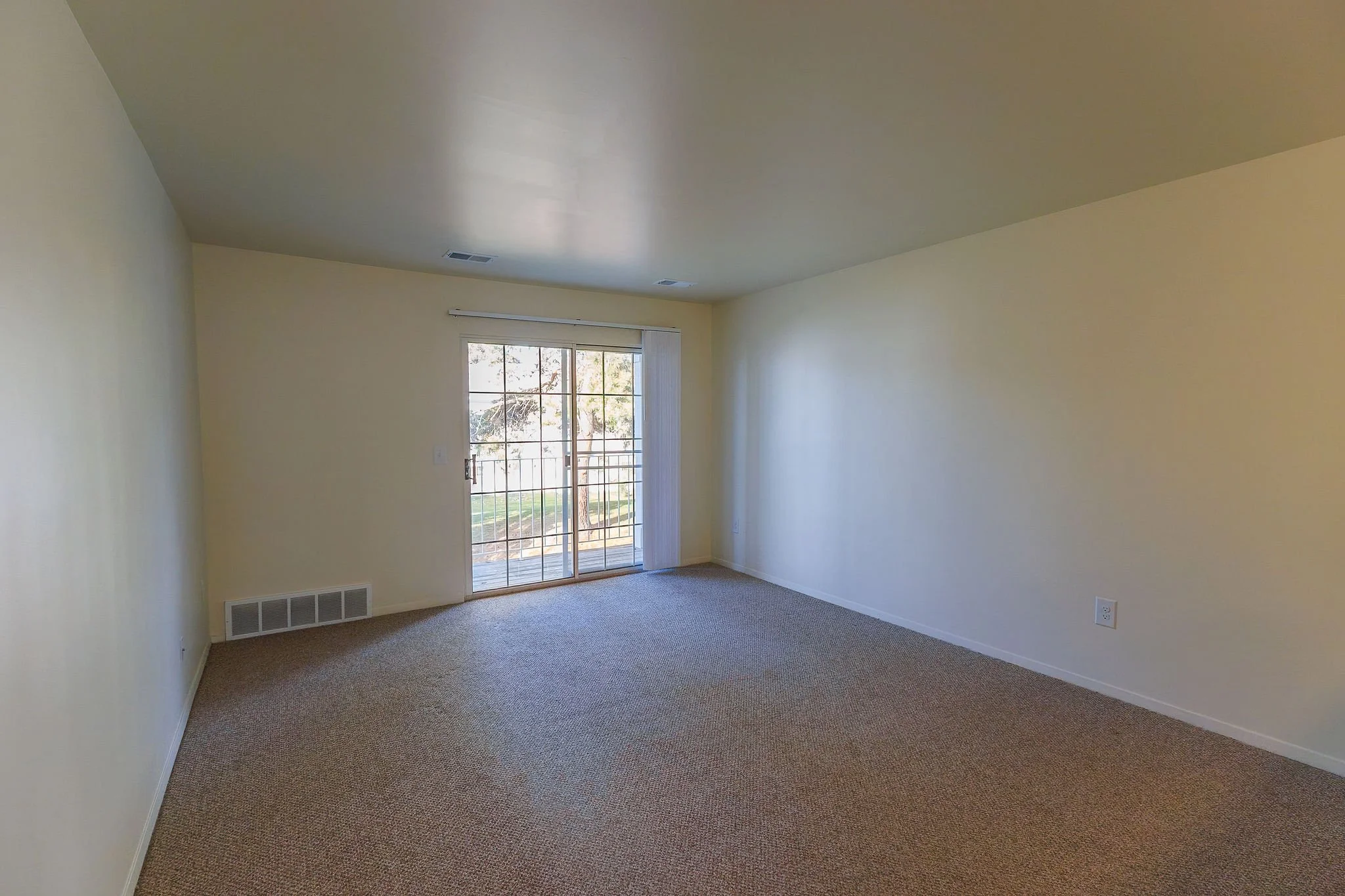 Empty living room with beige walls, brown carpet, sliding glass door leading to a patio, and a fenced yard outside.