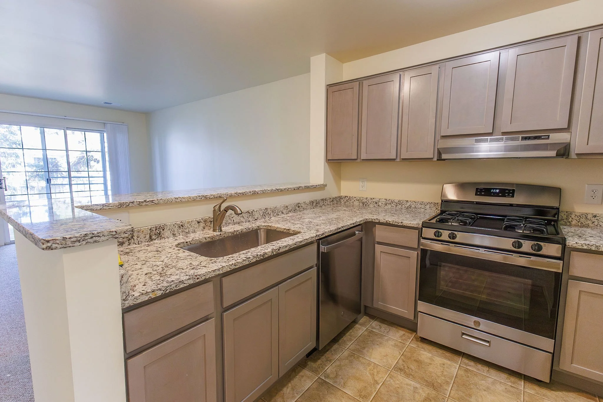 Kitchen with granite countertops, light wood cabinets, stainless steel stove, and a small under-sink dishwasher, overlooking a living area with a sliding glass door.