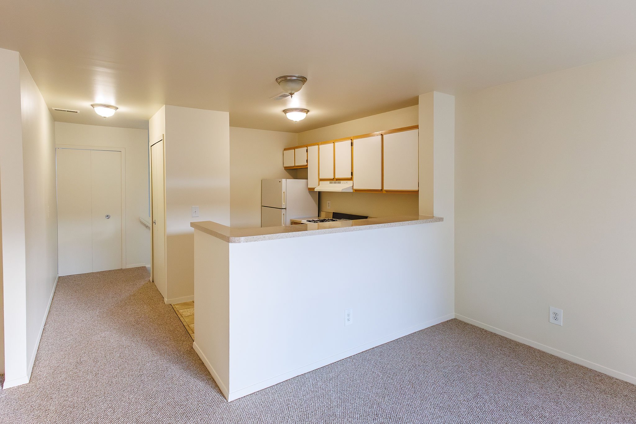 Empty apartment with a small kitchen that has white cabinets and a refrigerator, beige countertops, and a stove. Carpeted floor and plain white walls with ceiling lights.
