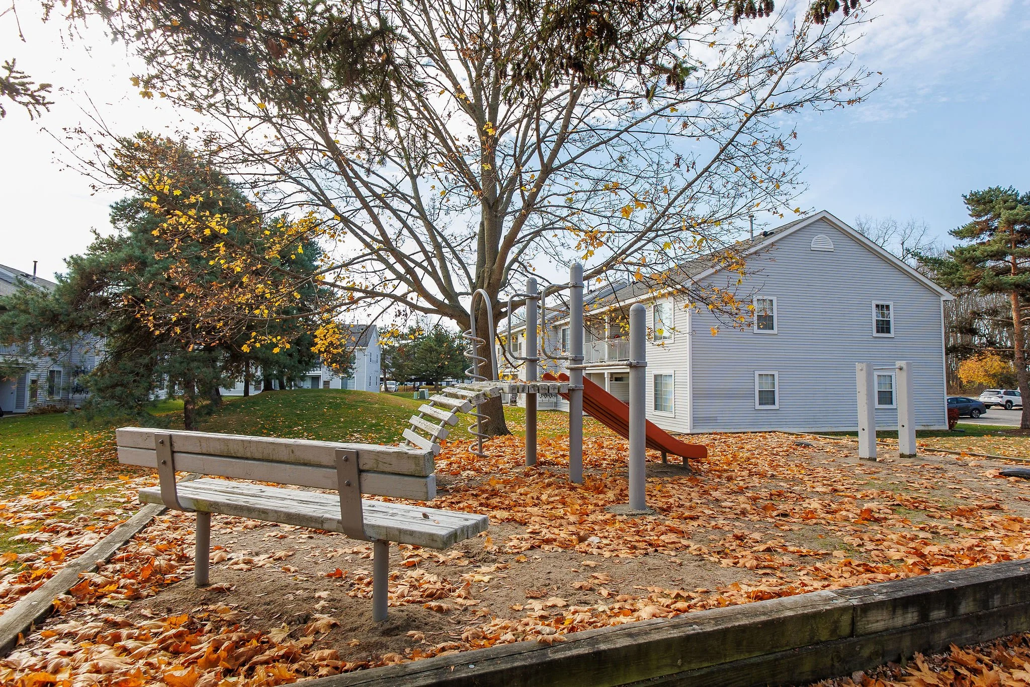 Empty playground with a slide, climbing equipment, and a wooden bench surrounded by fallen autumn leaves, with houses and trees in the background.