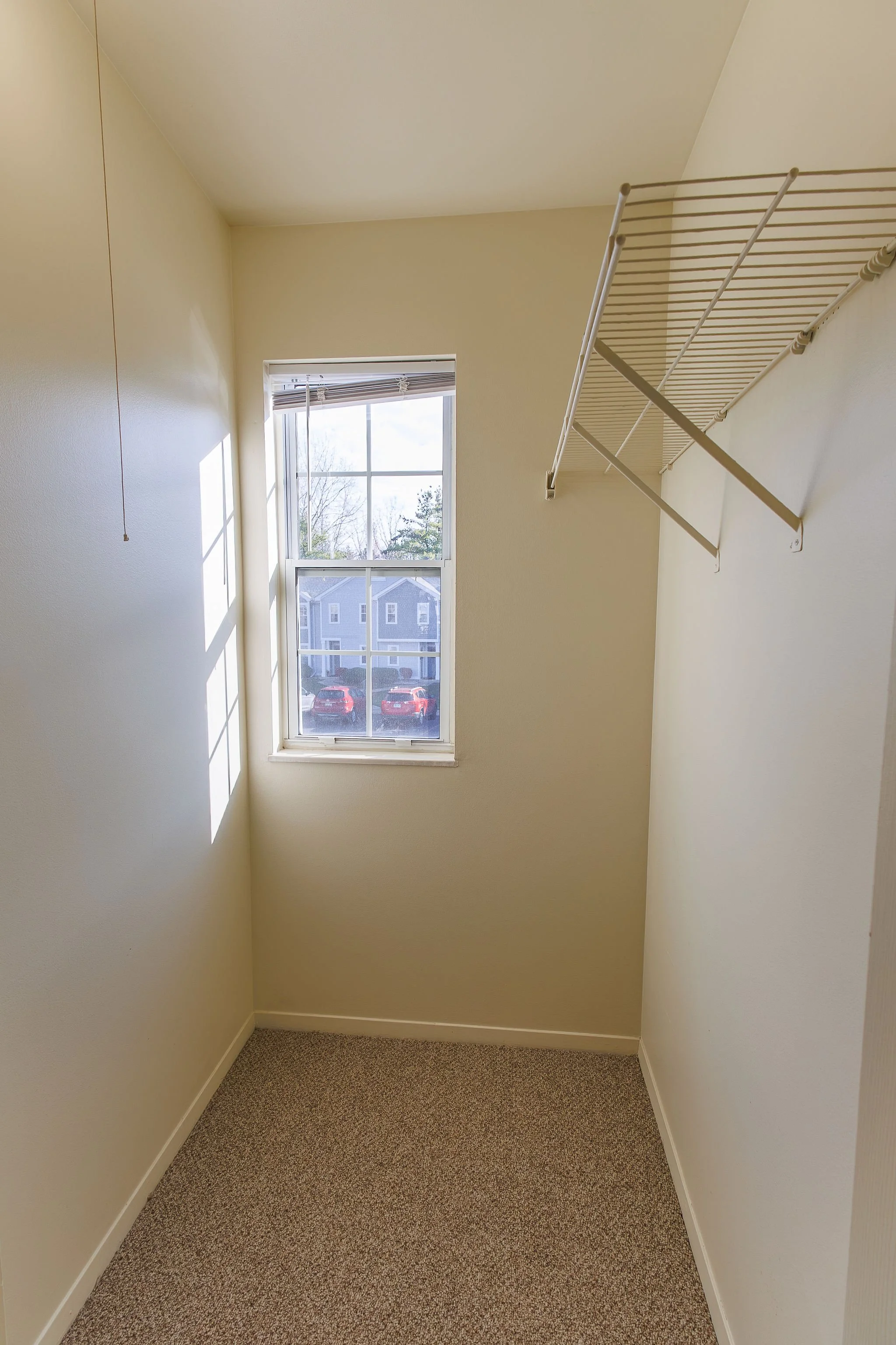 Empty walk-in closet with beige walls, a window letting in sunlight, and a white metal shelves mounted on the right wall.