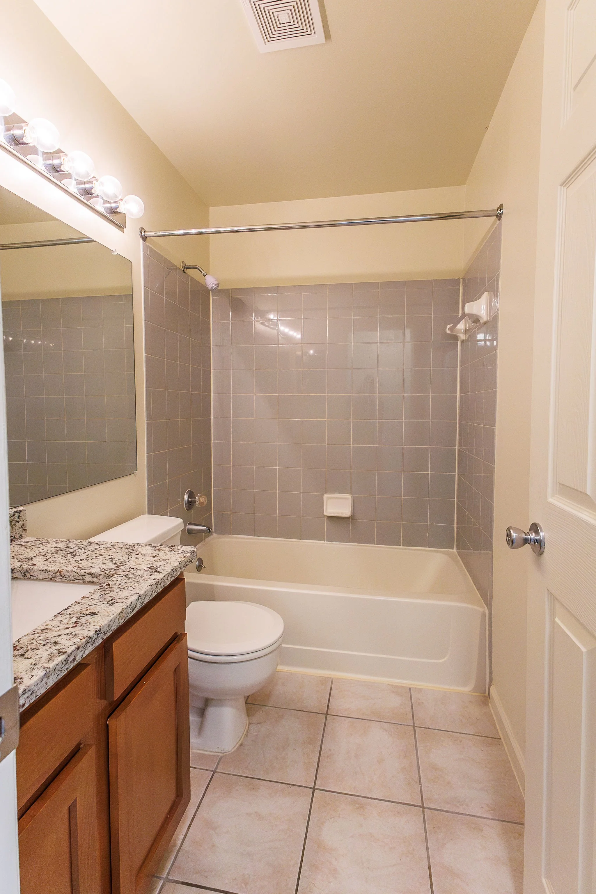 Small bathroom with a bathtub and shower with gray tiled walls, a white toilet, a wooden cabinet with a granite countertop, and a large mirror with vanity lights.