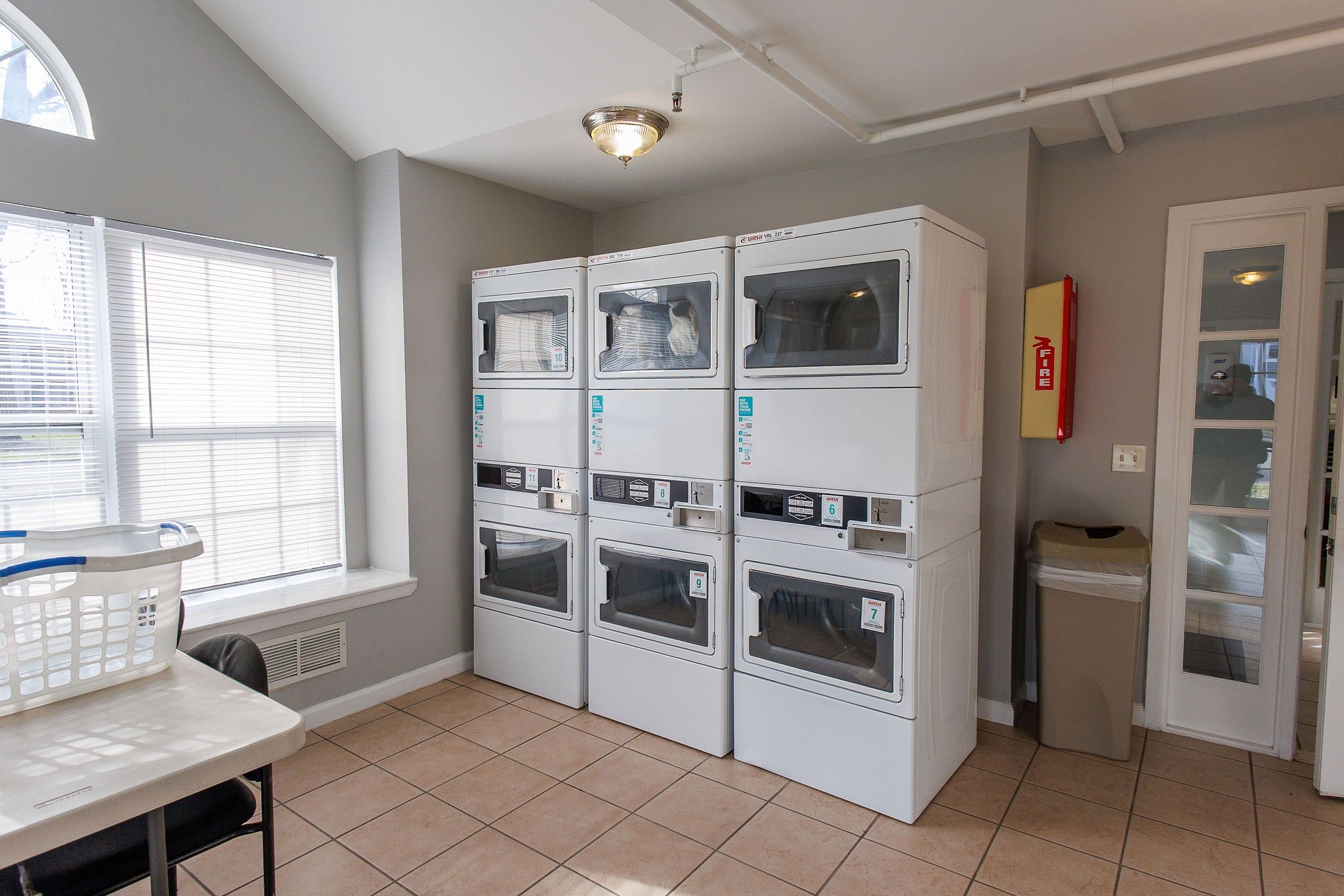 Laundry room with three stacked washers and dryers, a window with blinds, a table with laundry basket, trash can, and fire extinguisher on the wall.